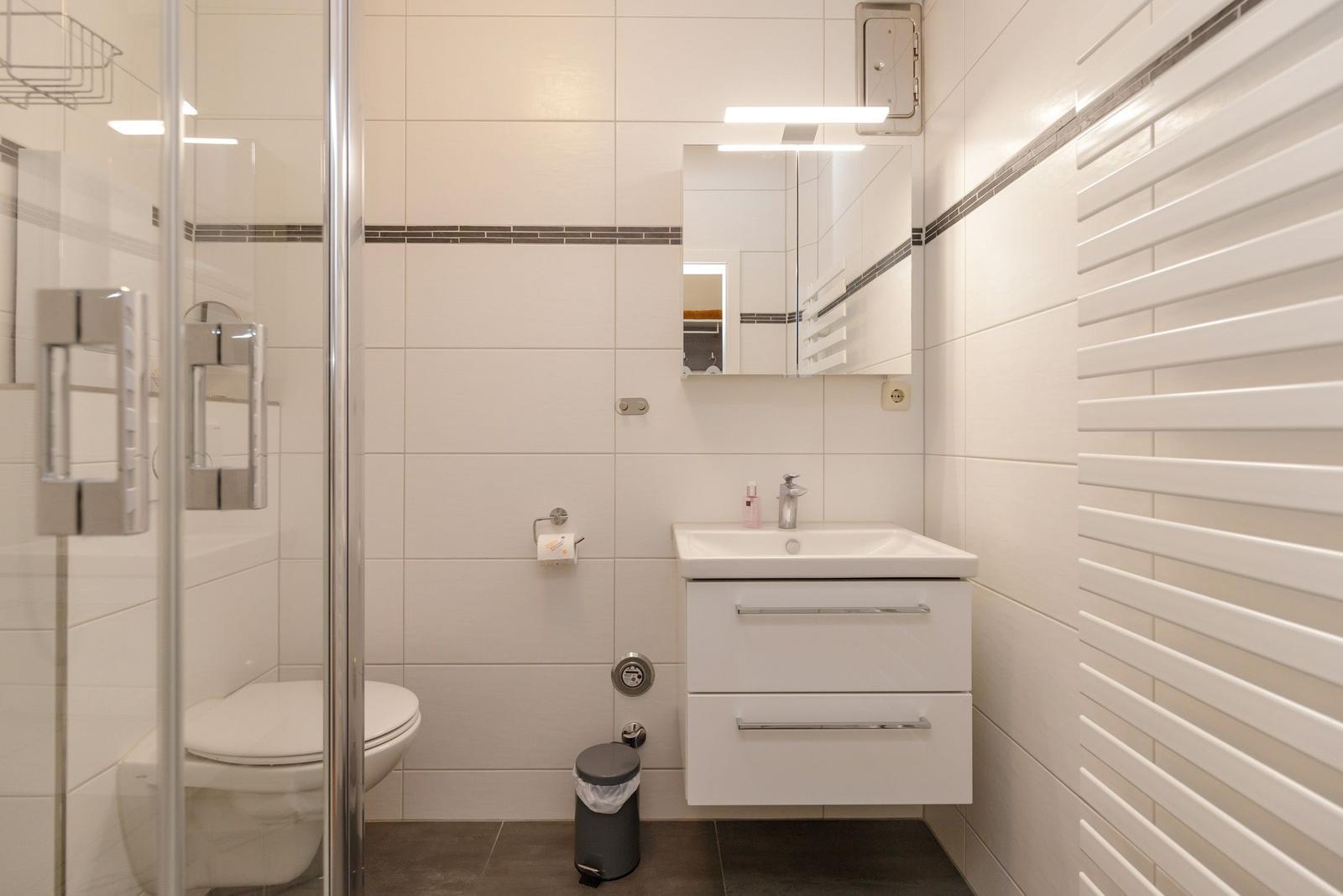 Badezimmer mit Dusche, Waschbecken und Toilette. Weiße Fliesen und Heizung.