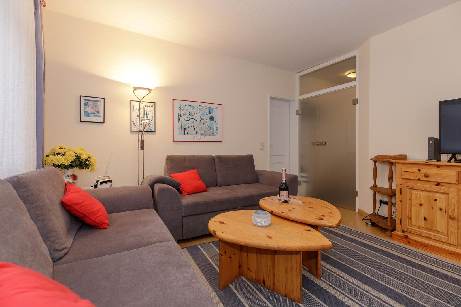 Wohnzimmer mit grauem Sofa, roten Kissen, Holztisch und Fernseher.