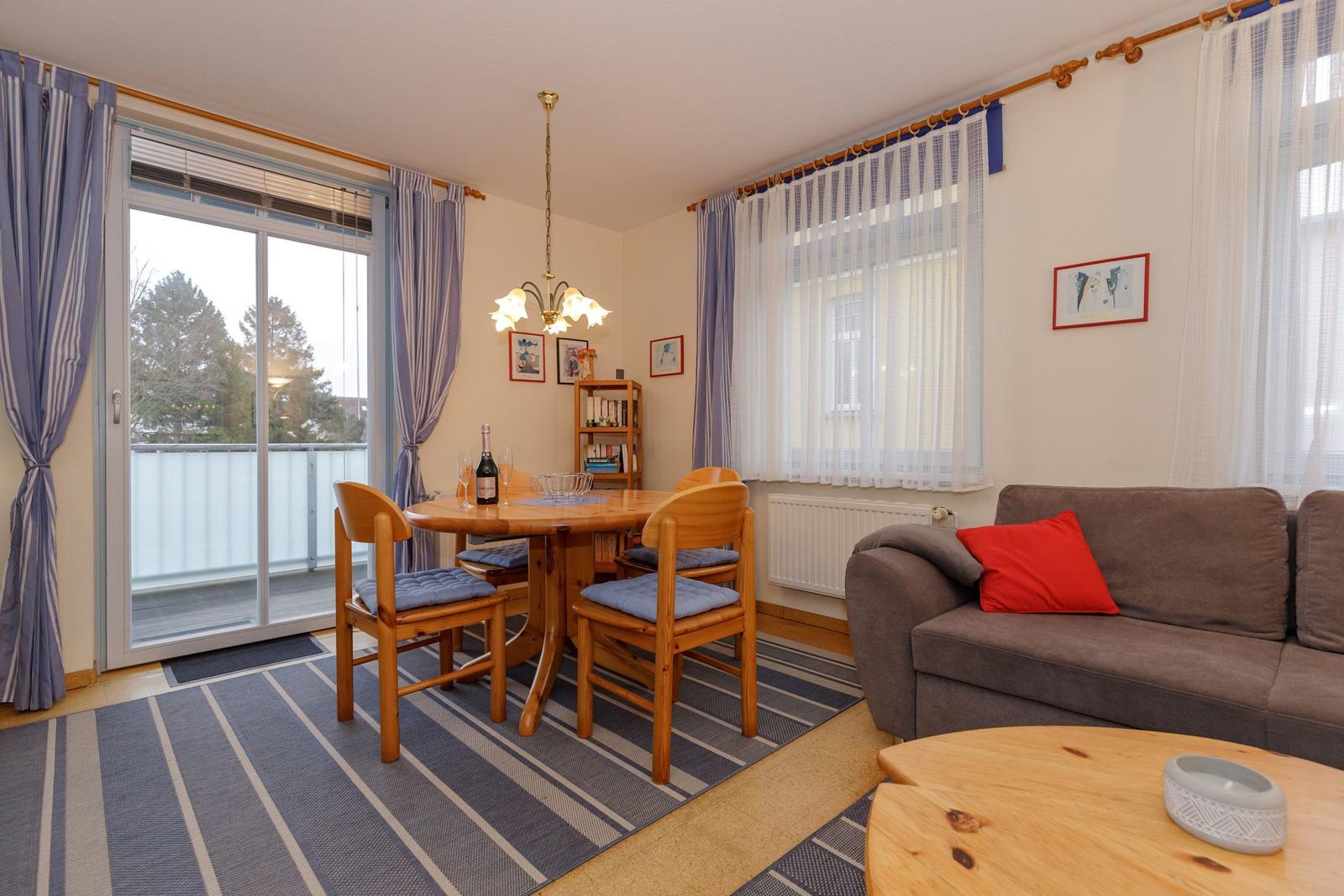 Wohn- und Essbereich mit Sofa, Tisch und Stühlen, Balkon und Fenster mit Vorhängen.