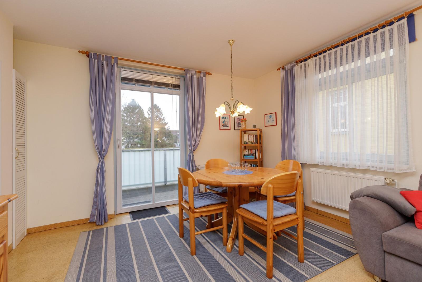 Esszimmer mit Holztisch, Stühlen und Balkontür. Sofa und Bücherregal sind sichtbar.