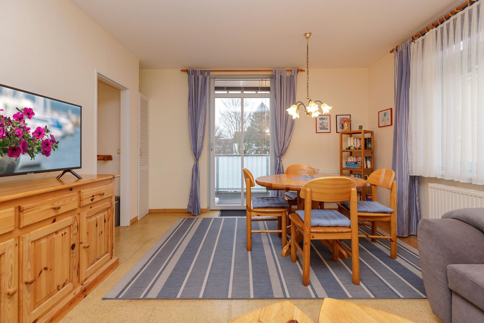 Wohn- und Essbereich mit Holztisch, Stühlen und Fernseher. Balkon mit Blick nach draußen.