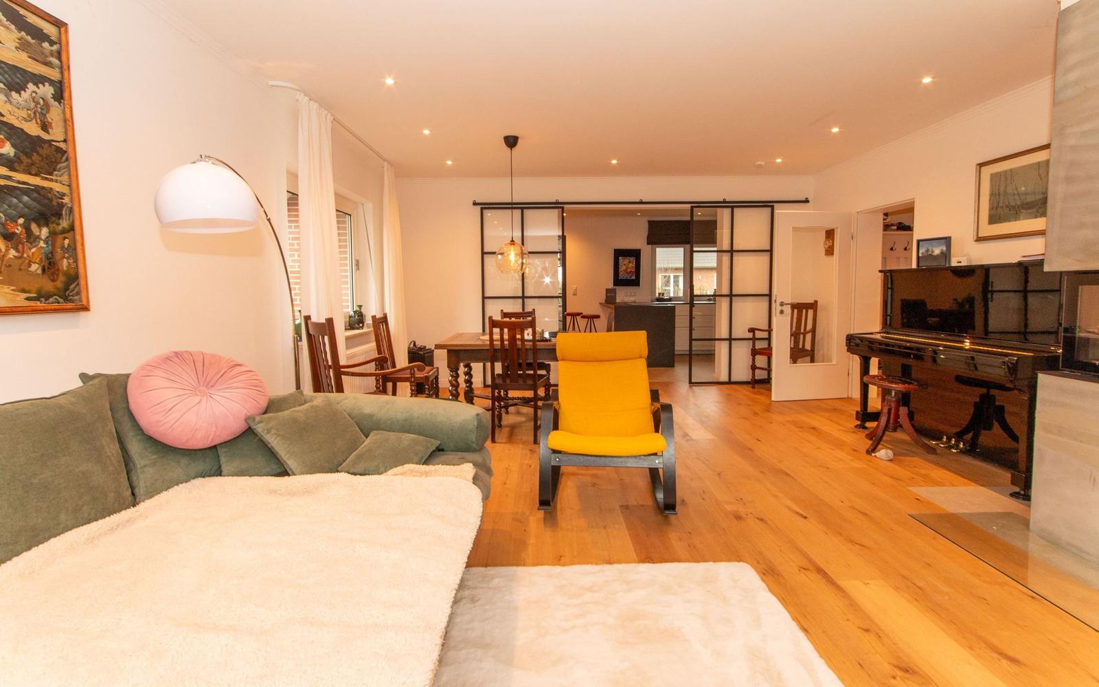 Wohnbereich mit Sofa, gelber Sessel und Klavier. Holzboden und Essbereich im Hintergrund.