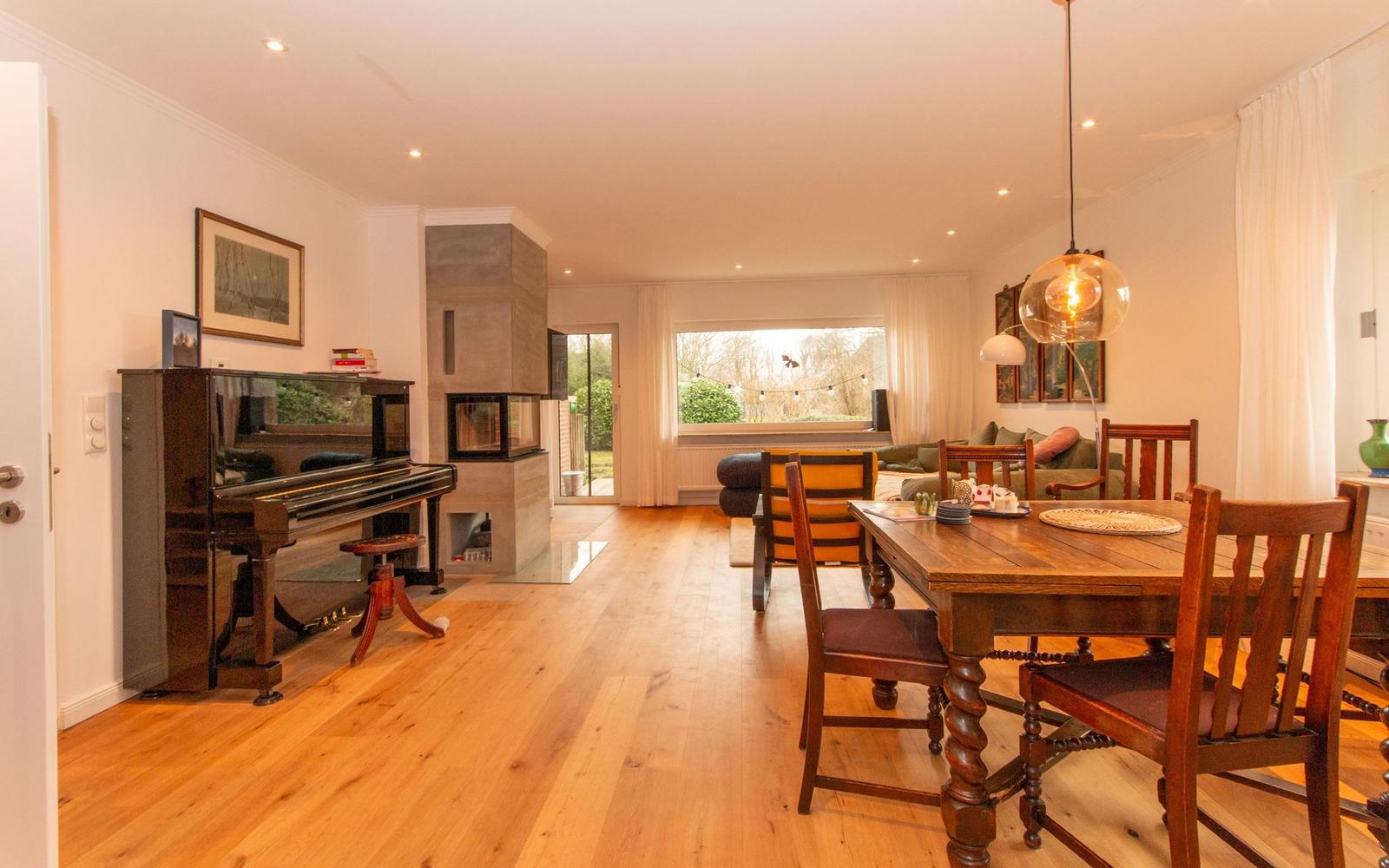 Ein Esszimmer mit Holztisch und Stühlen. Im Hintergrund befindet sich ein Wohnbereich mit Klavier und Kamin.