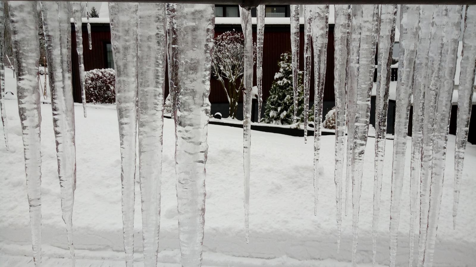 Eisstürze hängen von einem Dach, Schnee bedeckt den Boden und Bäume im Hintergrund.