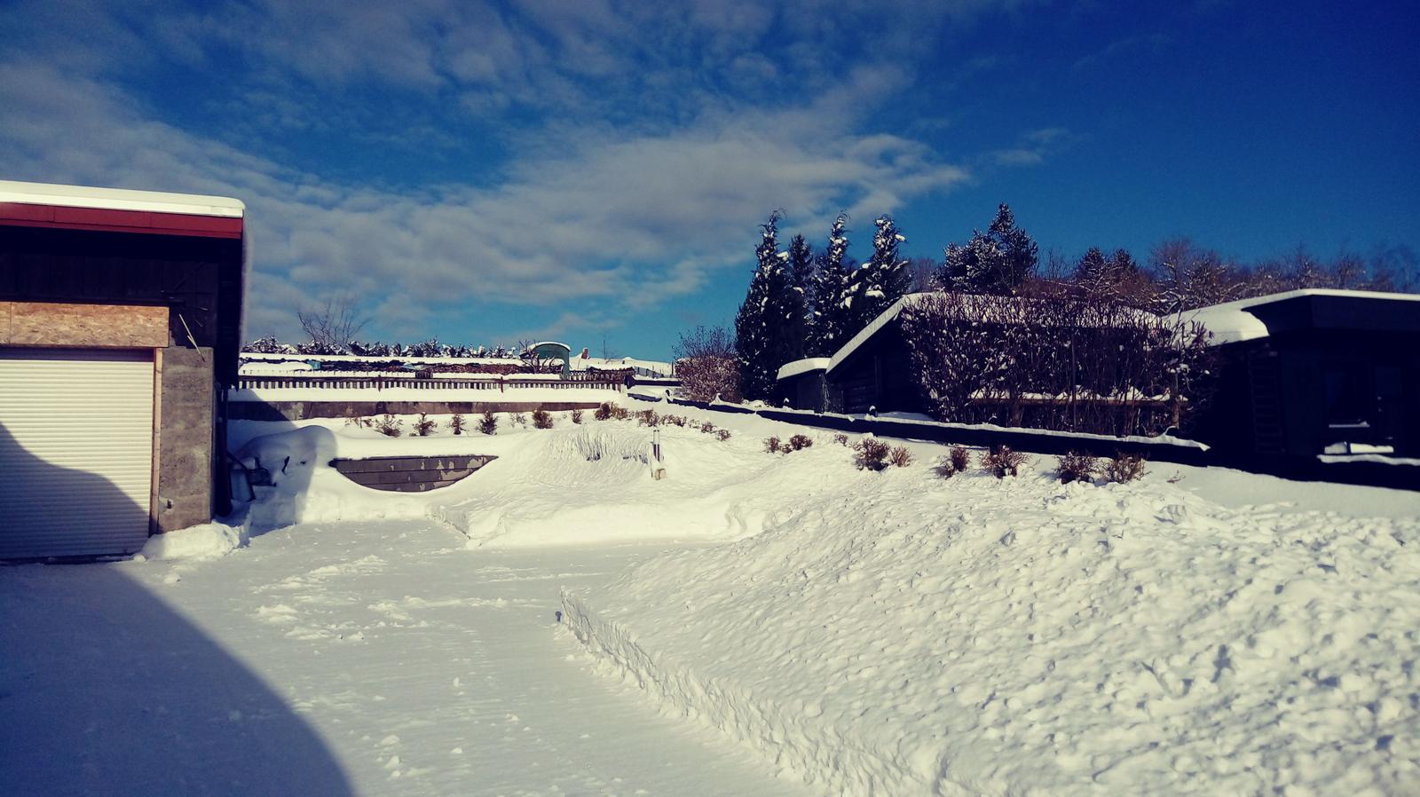 Schneebedeckter Außenbereich mit Garage und Gartenhaus unter blauem Himmel.
