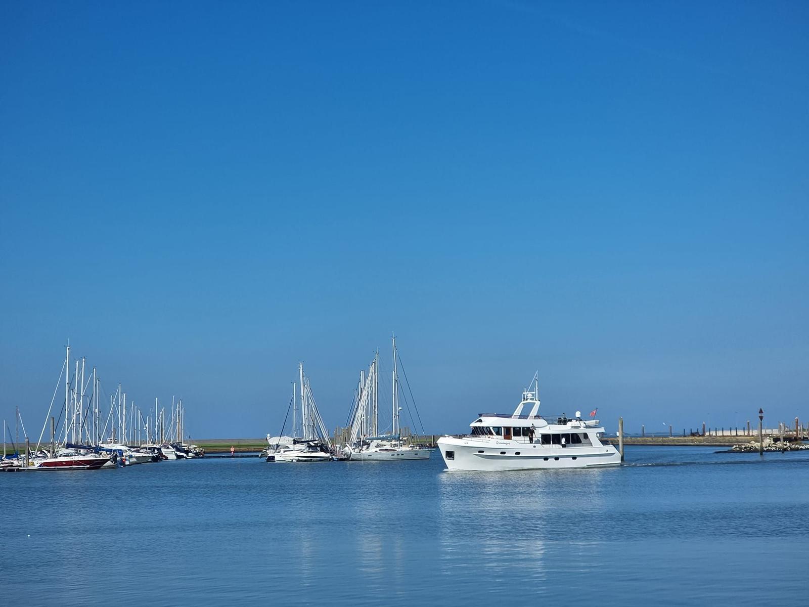 Ein weißes Motorboot fährt durch einen Hafen mit Segelyachten und klarem Himmel.