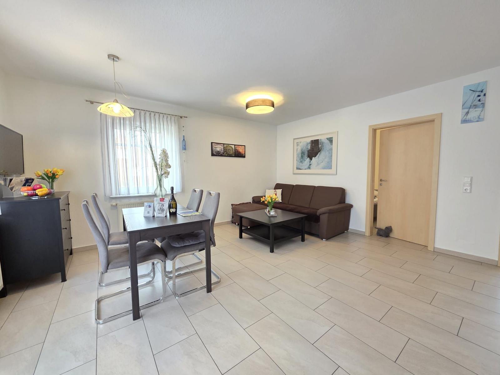 Wohn- und Essbereich mit Sofa, Tisch und Stühlen. Fenster, Wandbilder und Bodenfliesen.