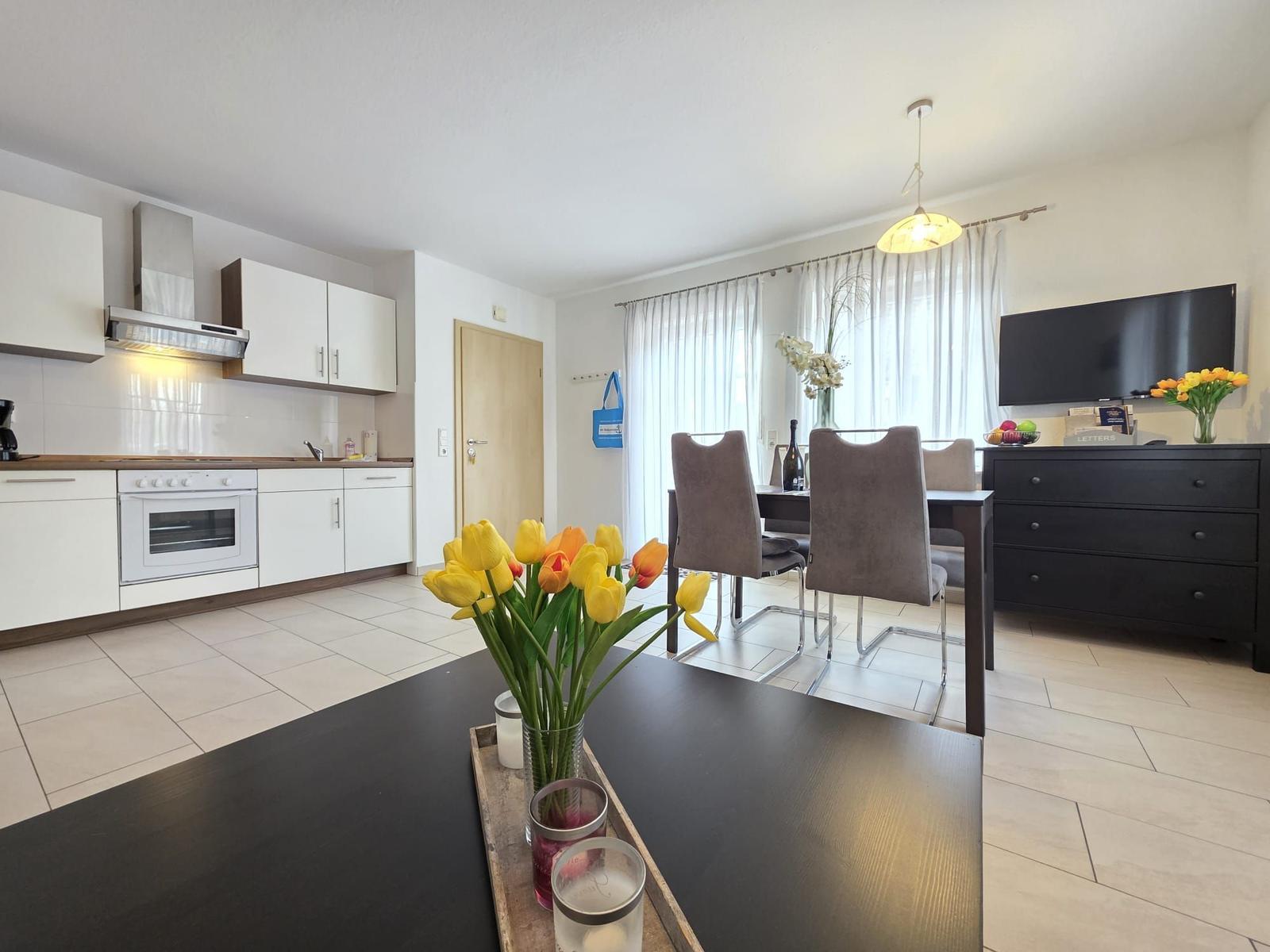 Küche, Essbereich und Wohnzimmer mit Fernseher und Blumen.