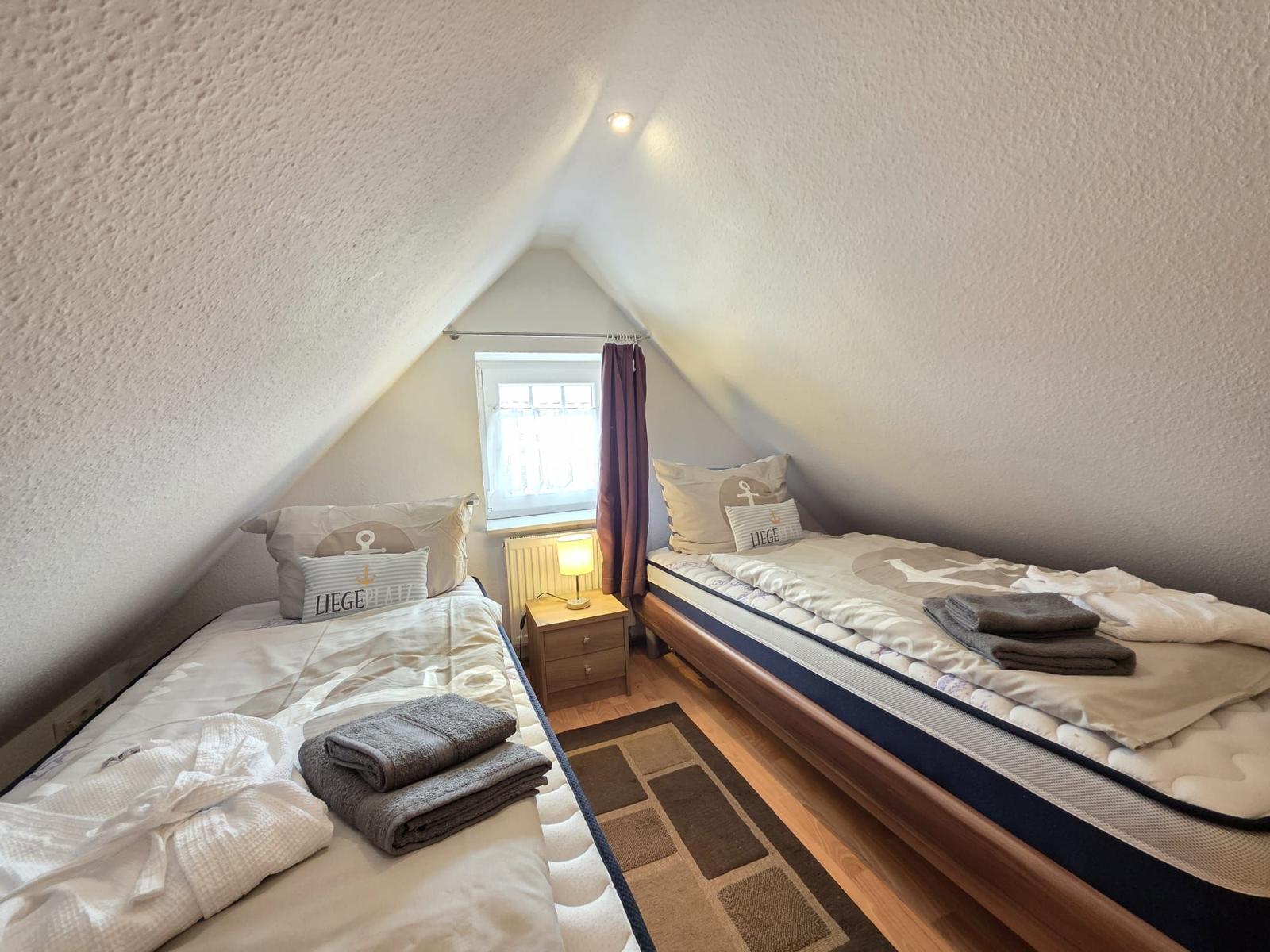 Two beds in attic room with window and nightstand.