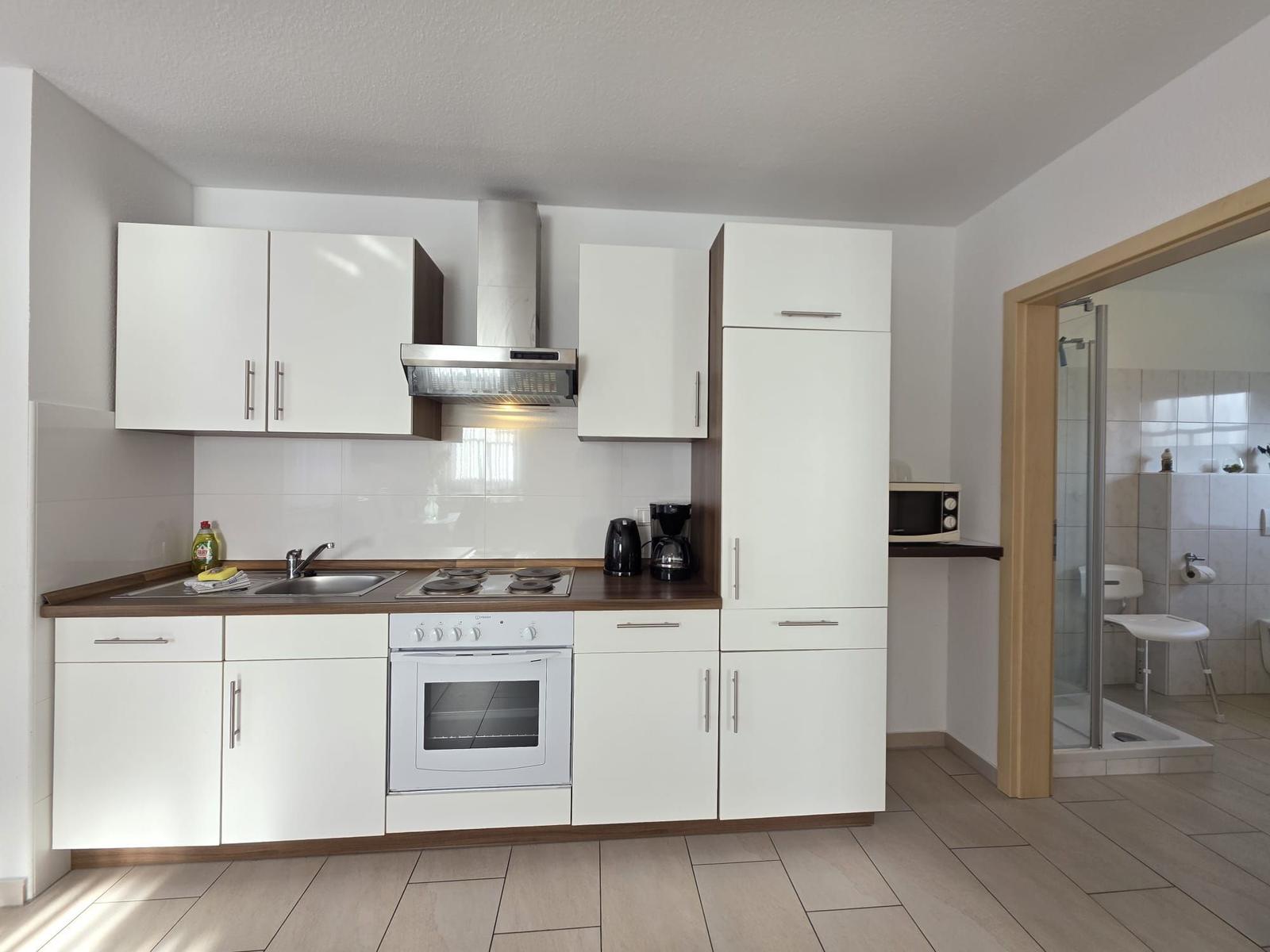 Kitchen with white cabinets, stove, and exhaust hood. Adjacent room is bathroom with shower.