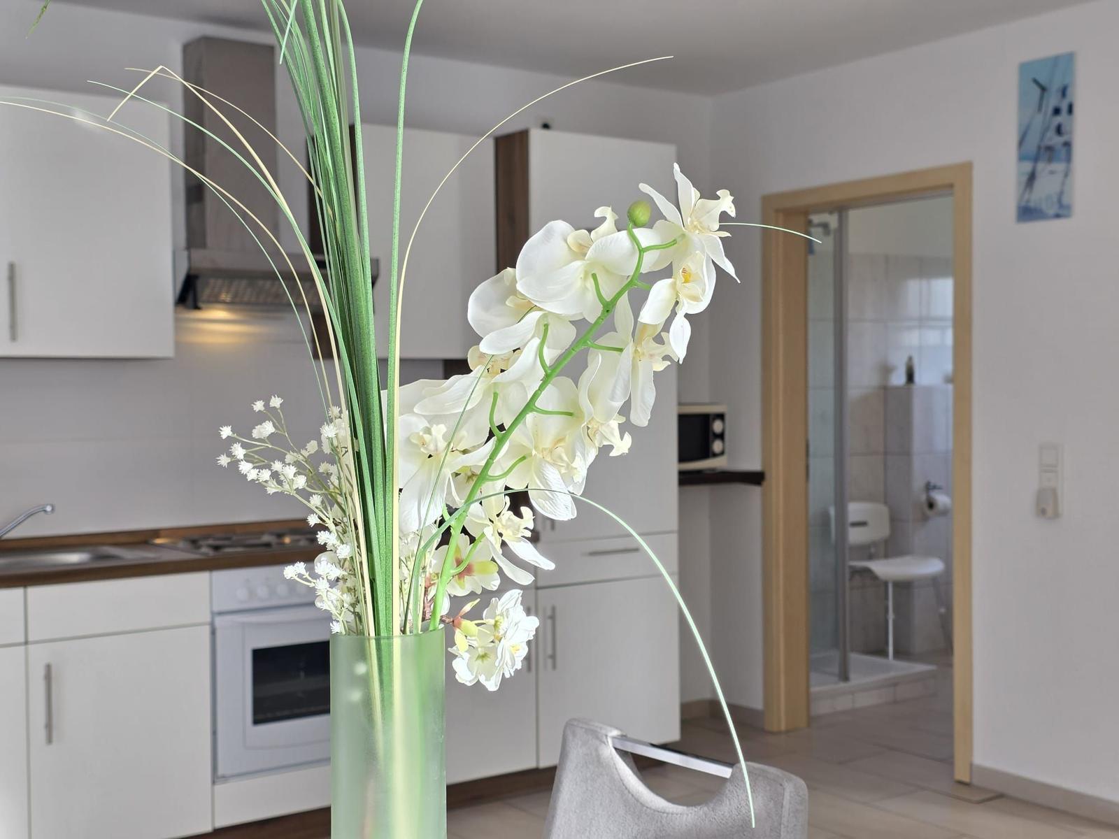 Kitchen with white cabinets and vase with white orchids. Passage to shower visible.