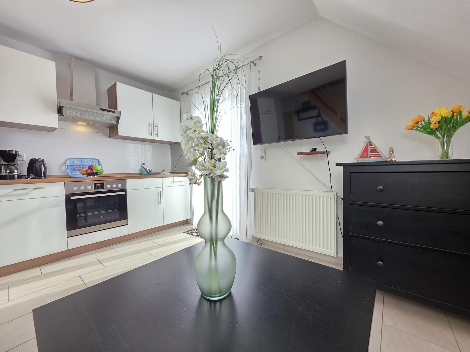 Kitchen with dining area, TV, and cabinet. Table with flower vase in foreground.