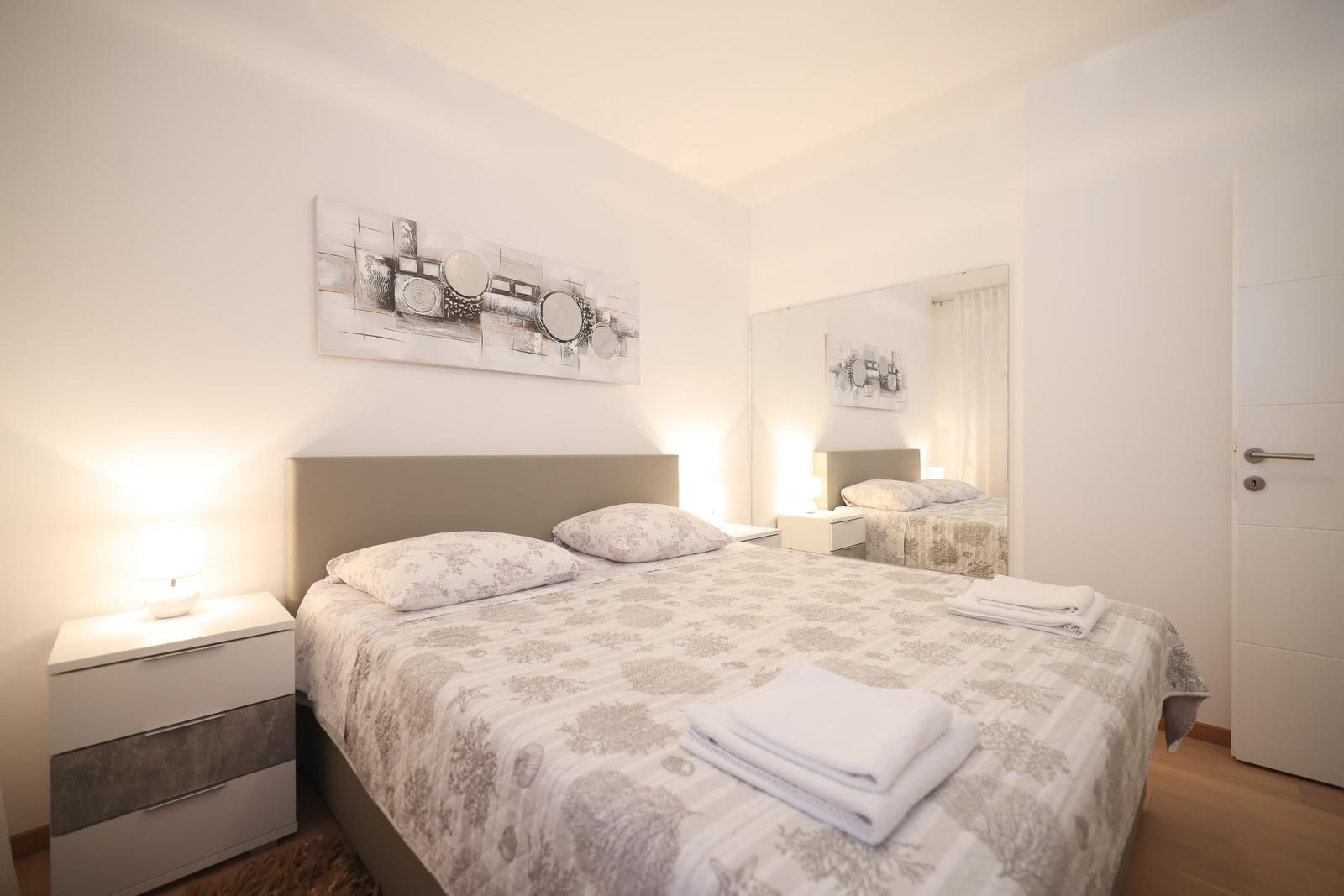 Schlafzimmer mit Doppelbett, Nachttisch und Wandbild. Spiegel reflektiert das Zimmer.