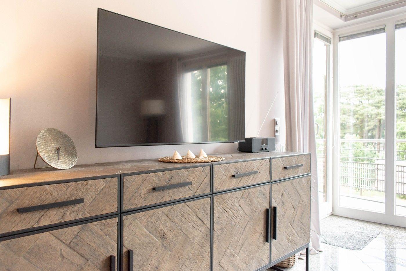 Flachbildfernseher über einem Holzschrank mit Schubladen und Türen. Durchgang zur Terrasse mit Blick auf Grün.