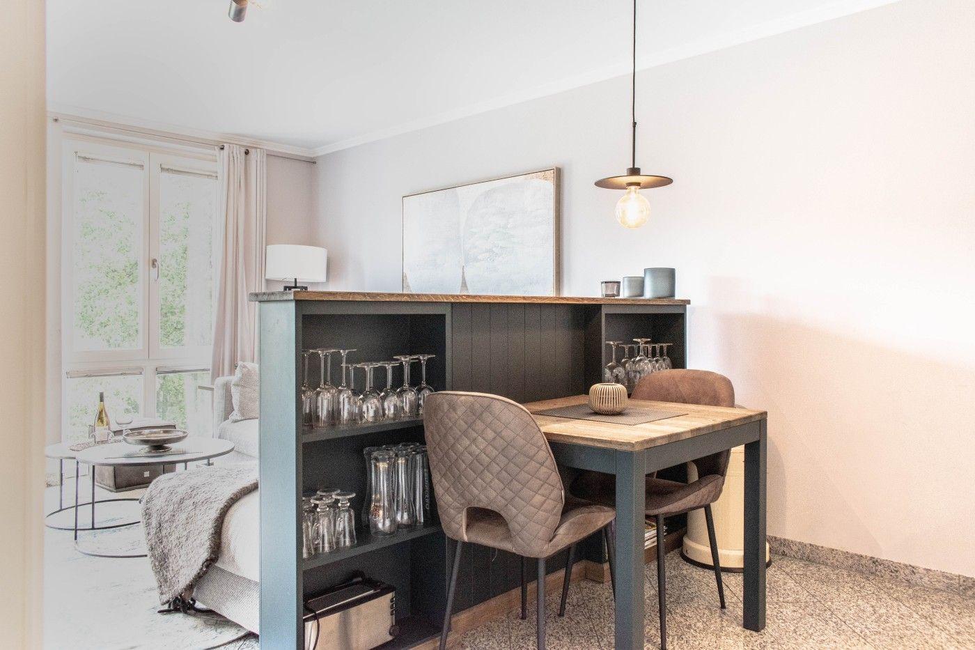 Essbereich mit Tisch, Stühlen und Gläsern. Im Hintergrund Wohnbereich mit Fenster.