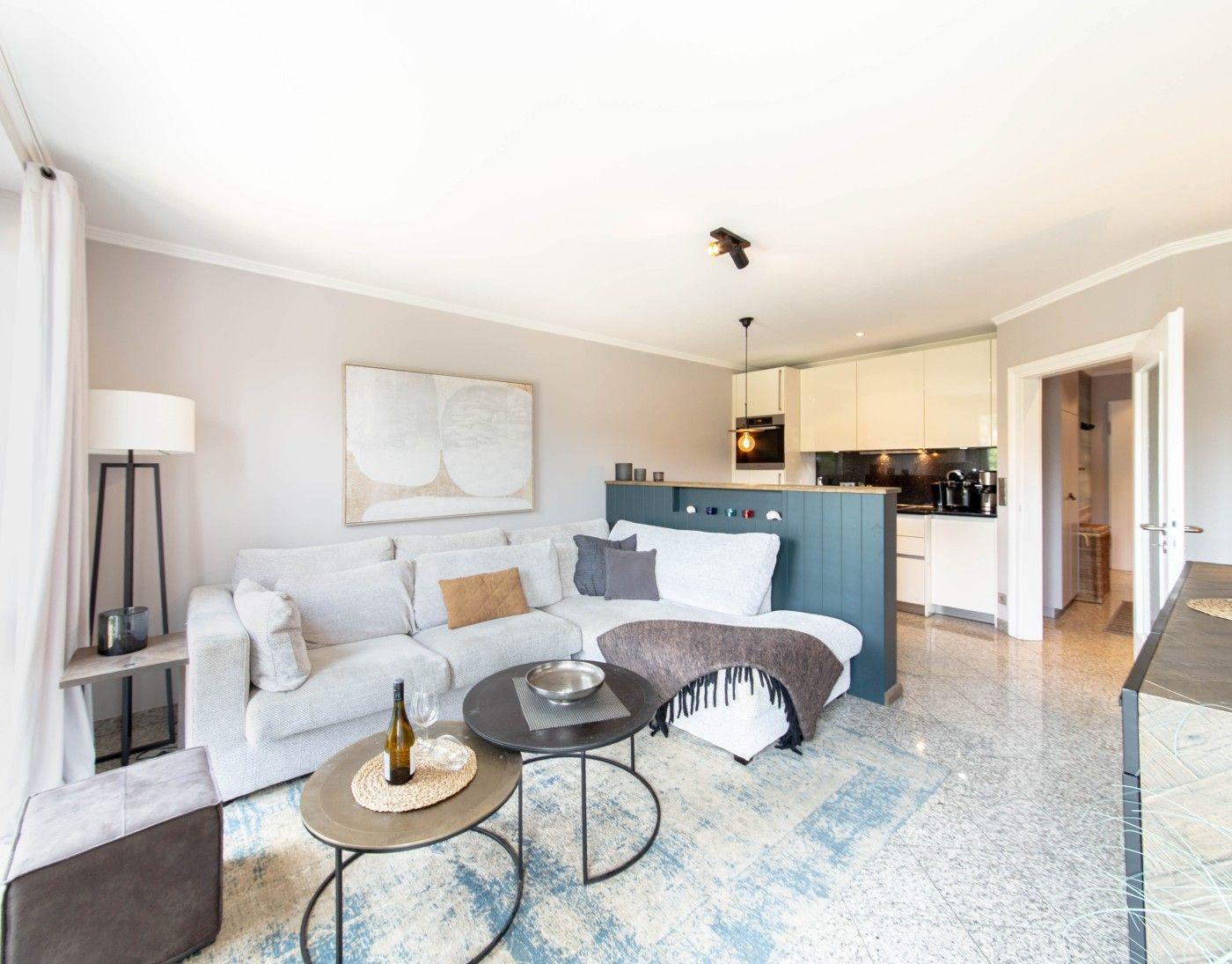 Wohnbereich mit Sofa, Tischen und Küche im Hintergrund.