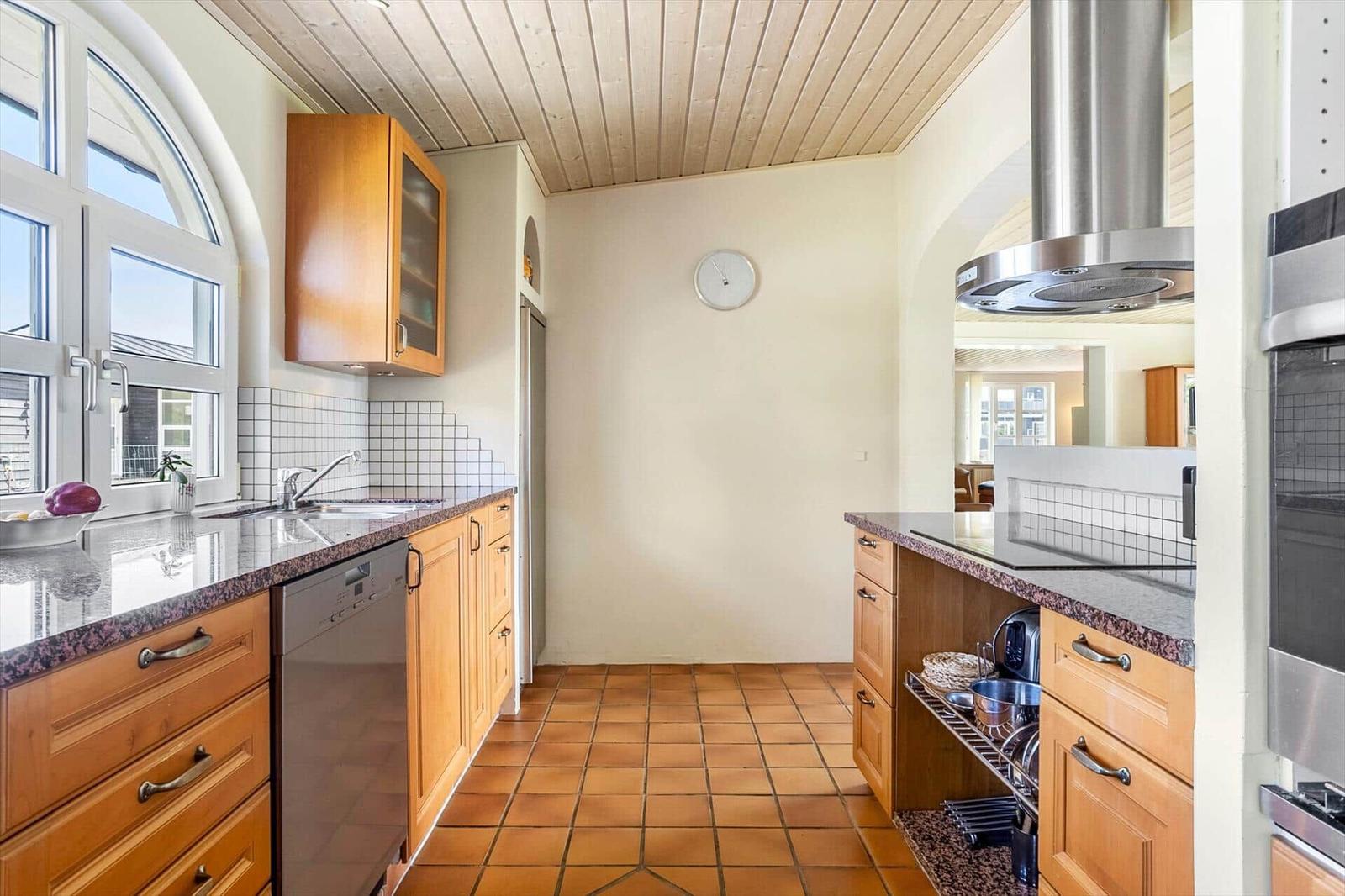 Kitchen with wooden cabinets, stone countertop, and terracotta floor. Window and range hood visible.