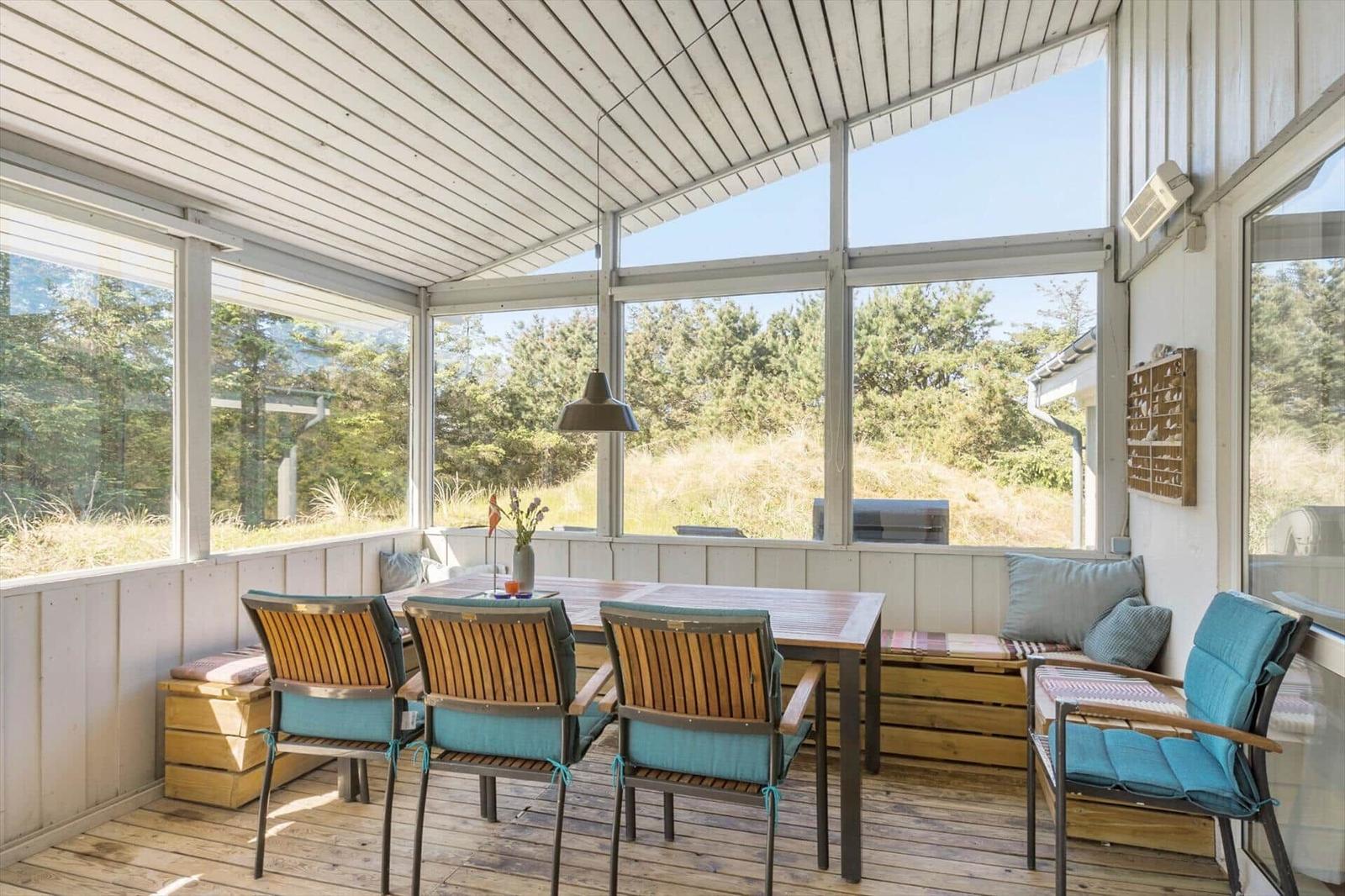 Terrace area with table, chairs, and bench. Large windows offer a view of the forest.