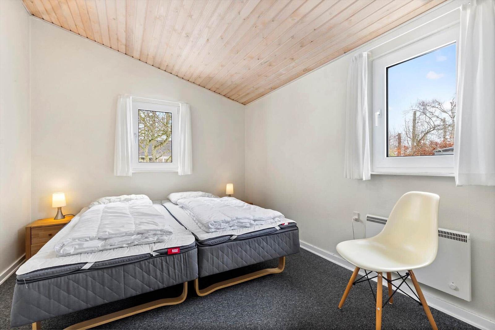 Two beds with mattresses, wooden ceiling, windows, and chair in room.
