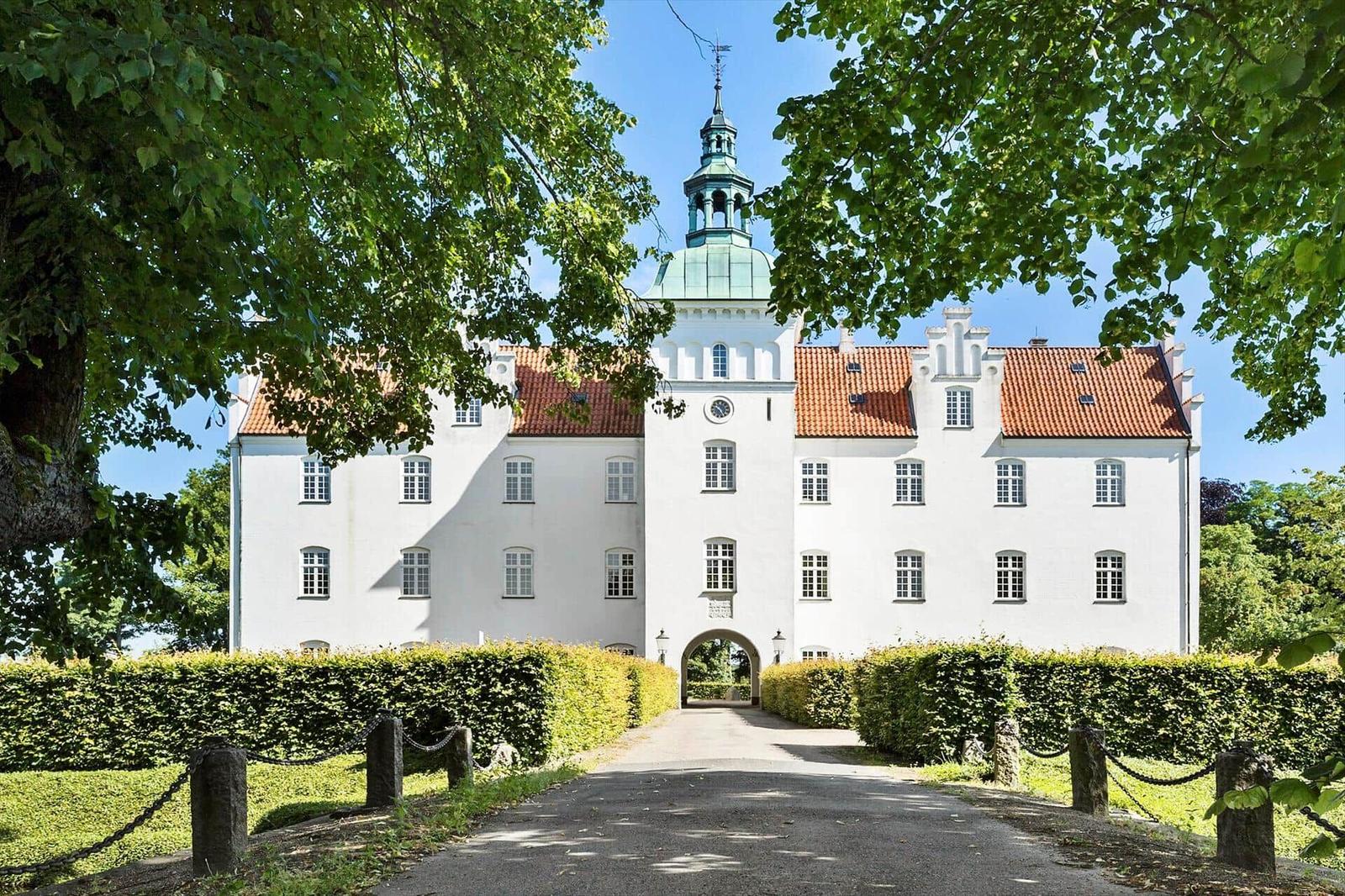 White castle with red roof and green dome, surrounded by trees and hedges.
