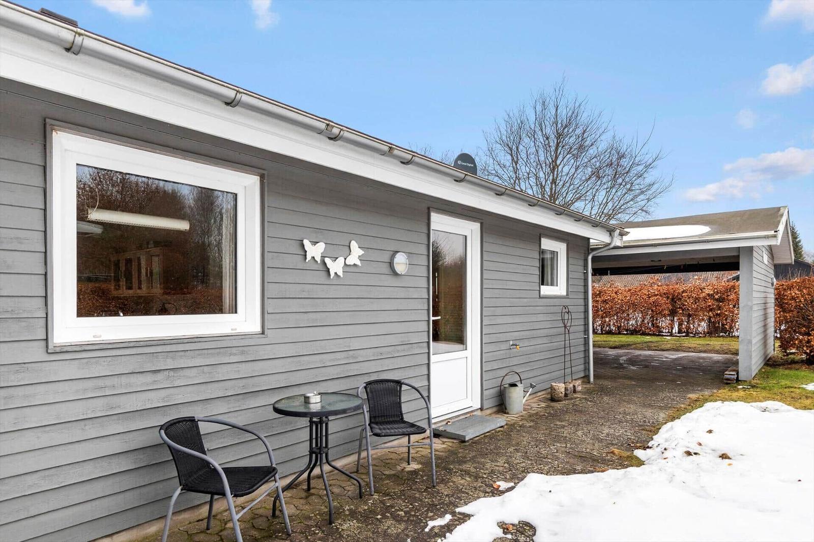 Gray wooden house with terrace, table, chairs, and snow on the ground.
