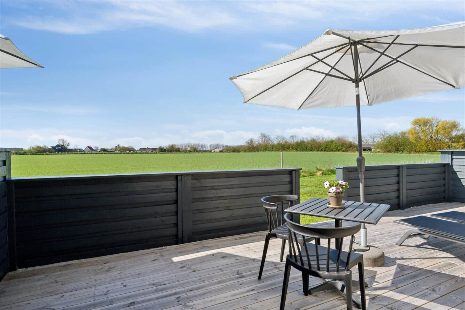 Terrasse mit Tisch, Stühlen und Sonnenschirm. Blick auf grünes Feld und Himmel.