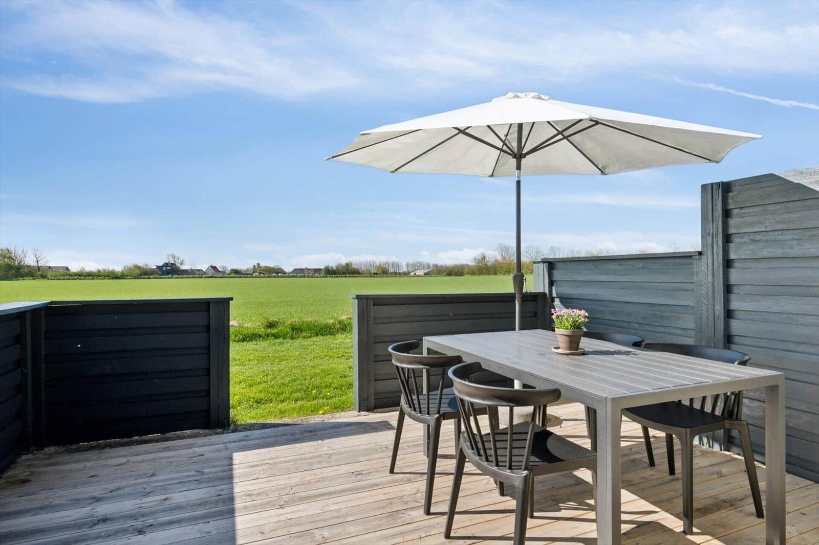 Terrasse mit Tisch, Stühlen und Sonnenschirm. Blick auf grünes Feld und Himmel.