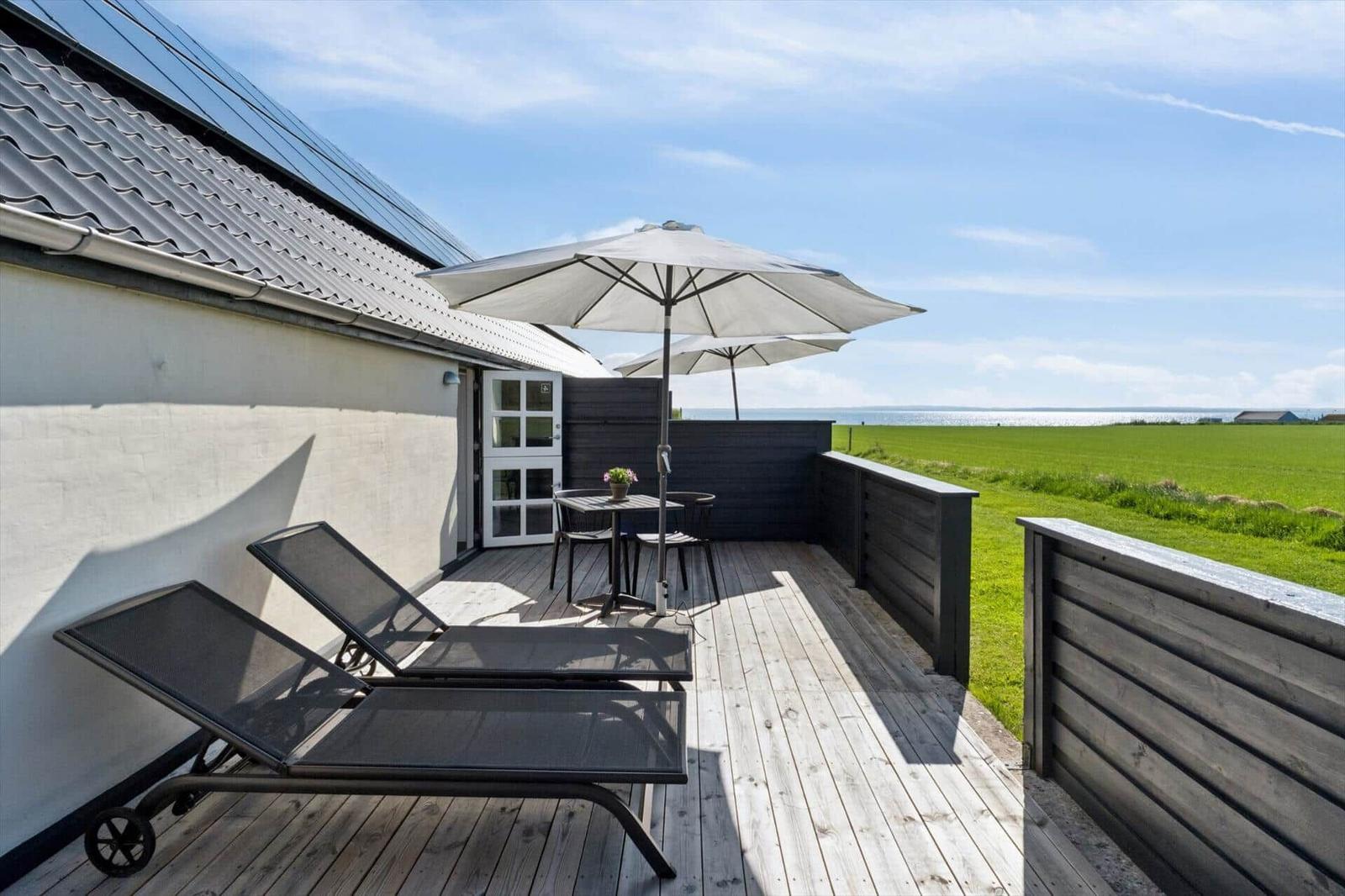 Terrasse mit Sonnenschirm, Tisch, Stühlen und Liegestühlen. Blick auf grünes Feld und Wasser.