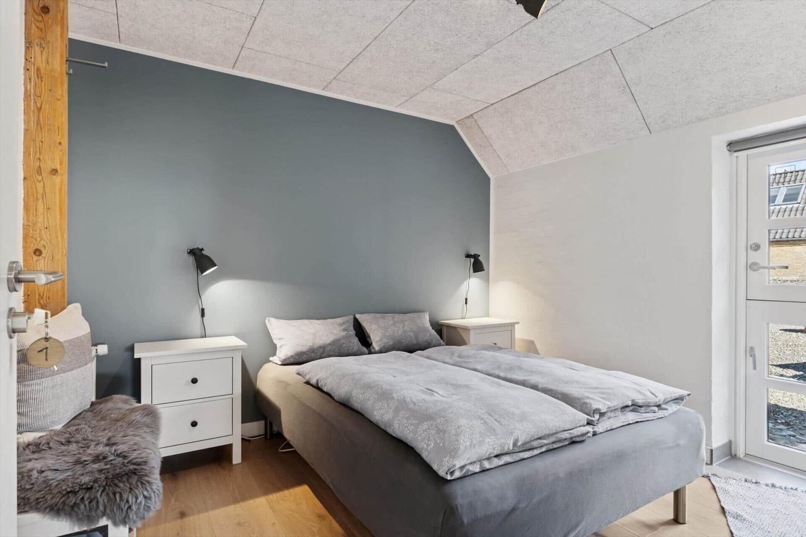Schlafzimmer mit Doppelbett, zwei Nachttischen und Fenster. Holzfußboden und graue Wand.