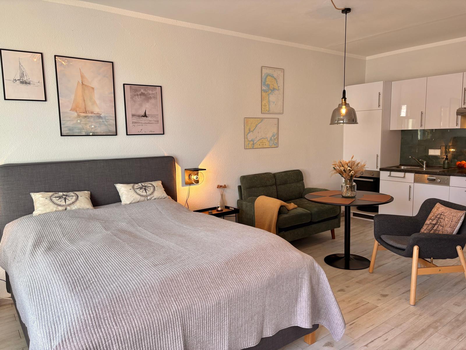 Schlafzimmer mit Bett, Sofa und Küchenbereich. Wandbilder zeigen Segelboote.