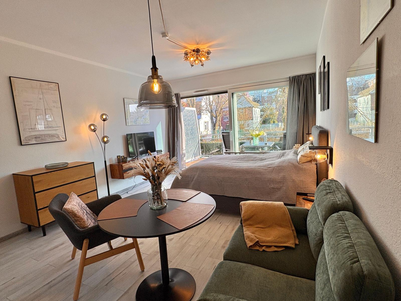 Ein Raum mit Schlafbereich, Wohnbereich und Essplatz. Große Fensterfront mit Blick auf Balkon.