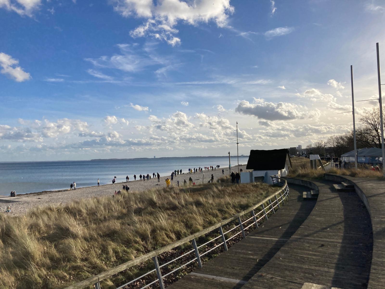 Holzweg entlang der Strandpromenade mit Dünen und Meer im Hintergrund.