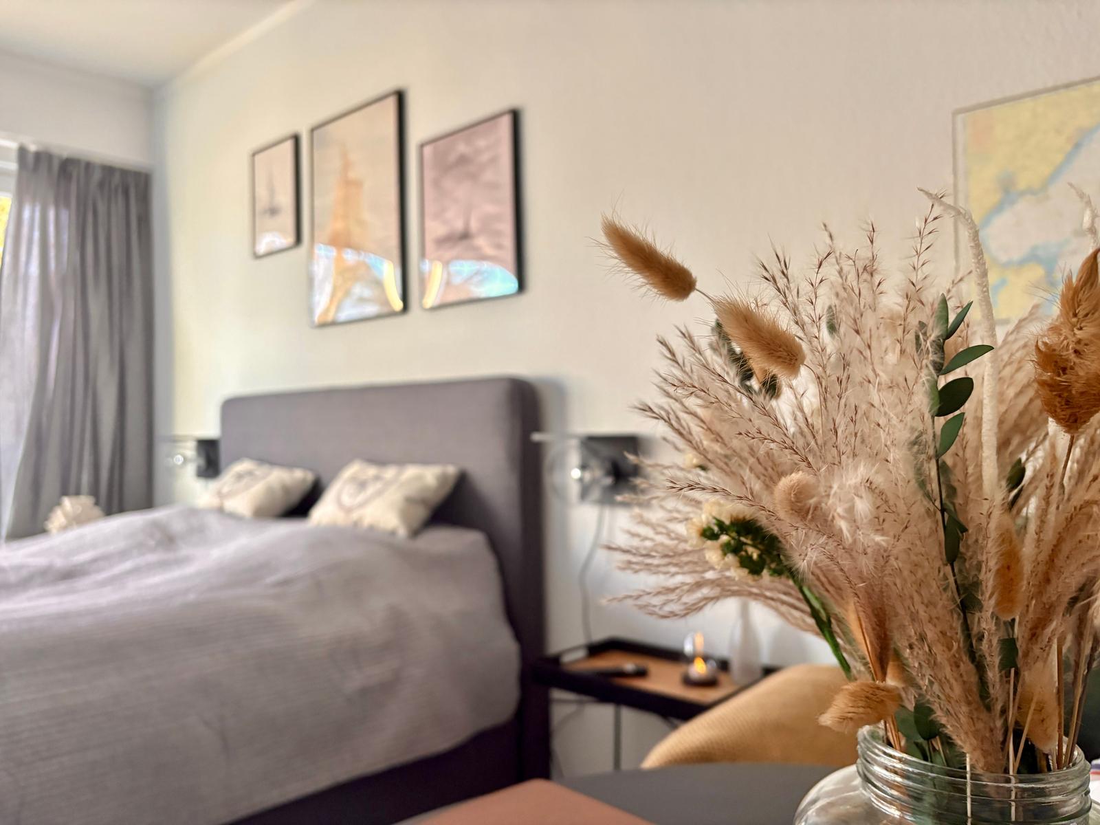 Schlafzimmer mit Bett, Wandbildern und Trockenblumen in Glasflasche.