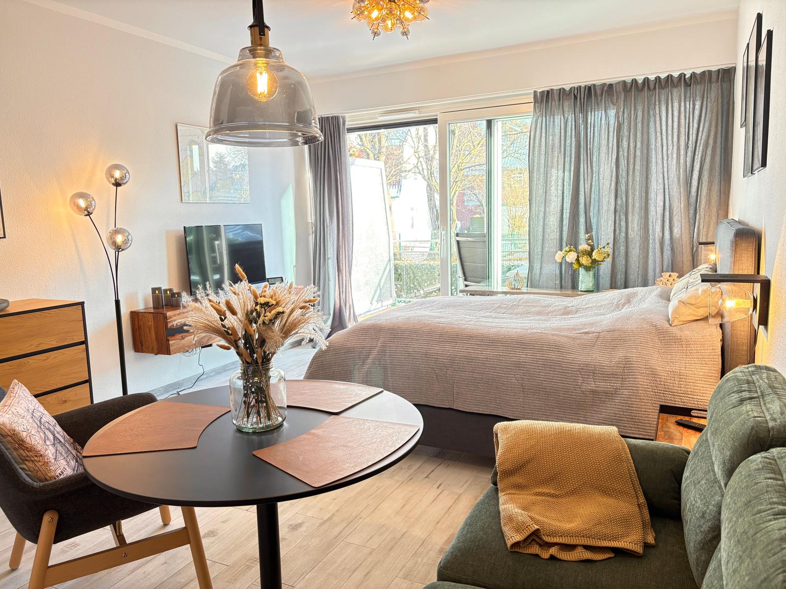 Ein Schlafzimmer mit Bett, Sofa, Tisch und Fernseher. Große Fenster mit Blick auf Balkon.