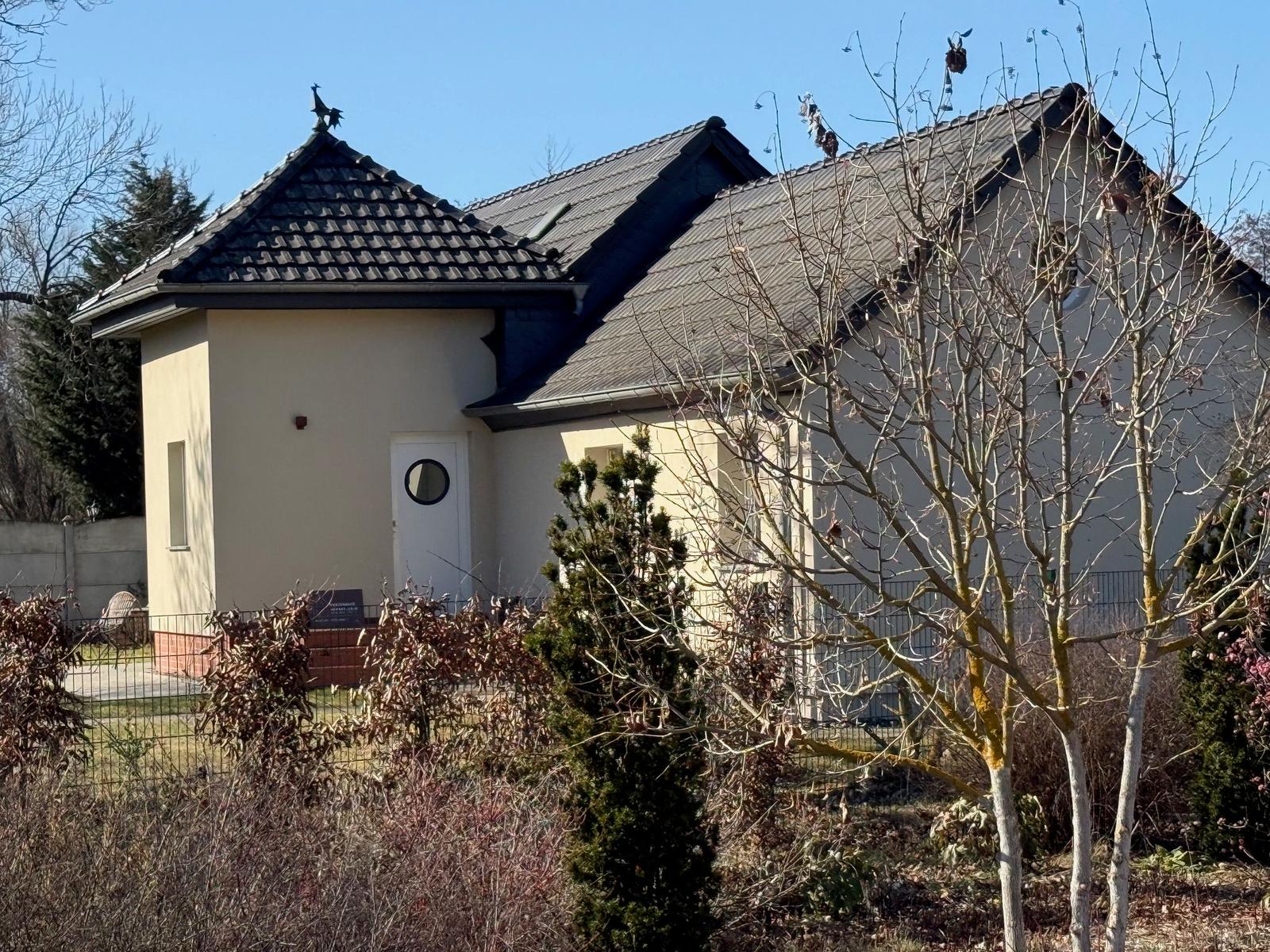 Ein Haus mit cremefarbener Fassade und dunklem Dach, umgeben von Bäumen und Sträuchern.