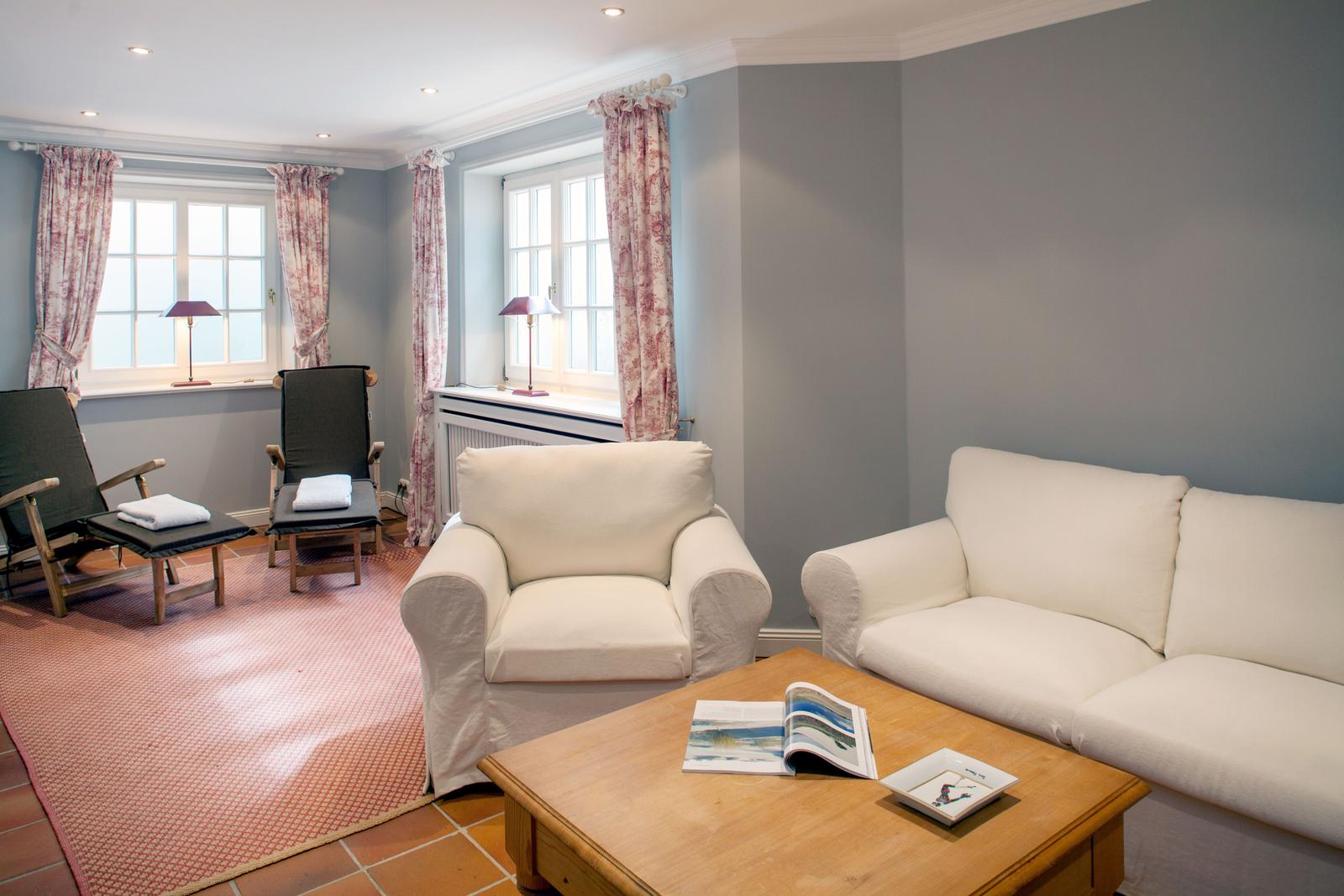 Wohnzimmer mit Sofa, Sessel, Tisch und Fenstern mit Vorhängen.