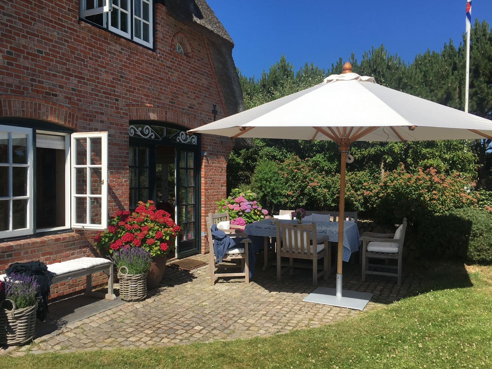 Terrasse mit Tisch, Stühlen und Sonnenschirm vor einem Backsteinhaus mit Blumen.