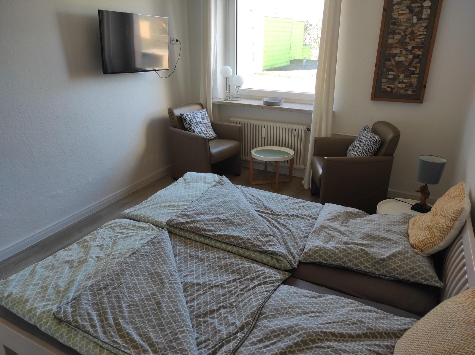 Schlafzimmer mit Bett, zwei Sesseln und Wandfernseher. Fenster mit Blick nach draußen.