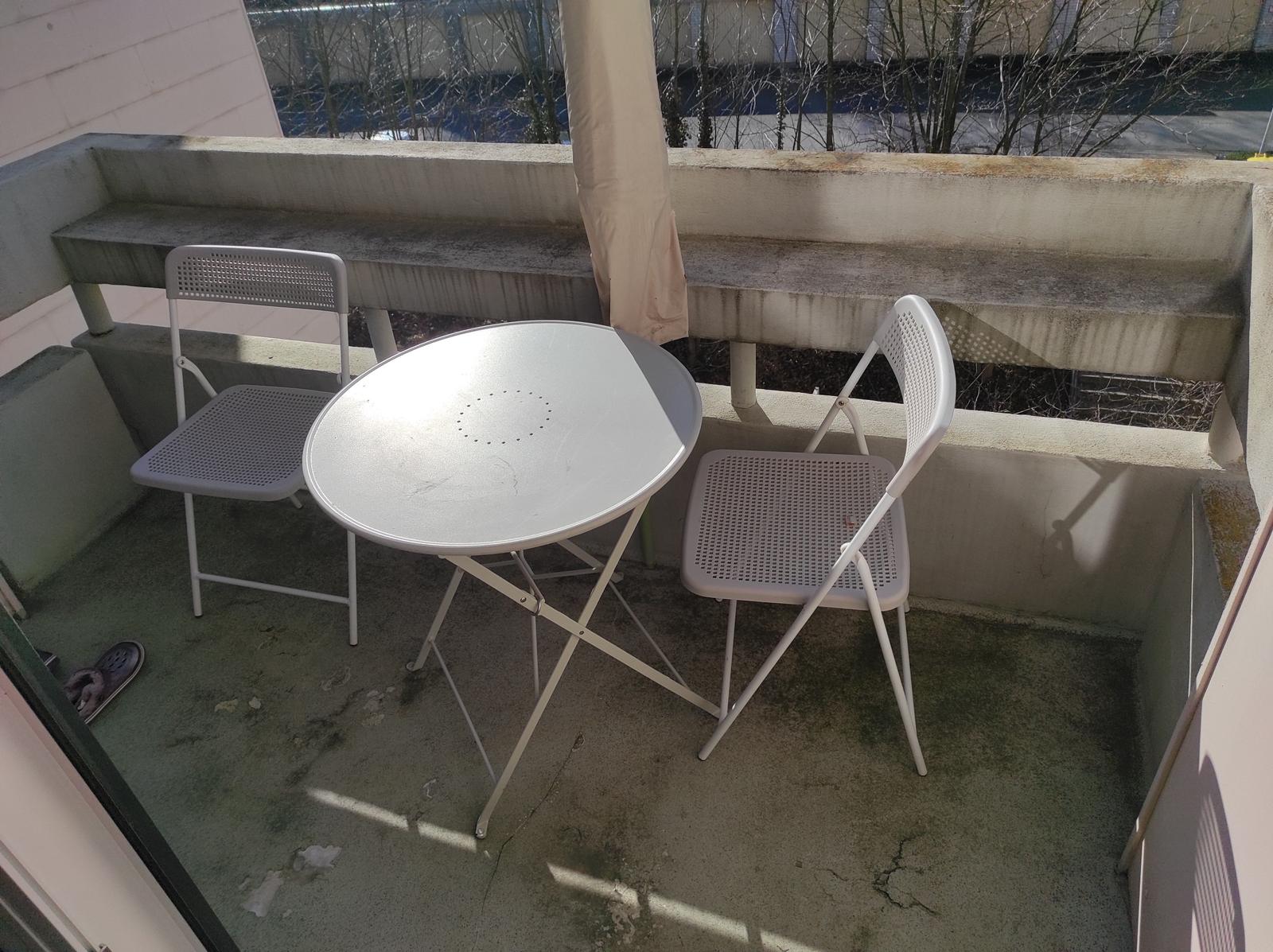 Balkon mit runden Tisch und zwei Stühlen, Hintergrund mit Bäumen und Gebäude.