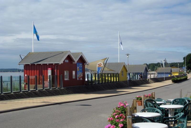 Strandpromenade mit bunten Hütten und Tischen. Blick auf Wasser und Flaggen.