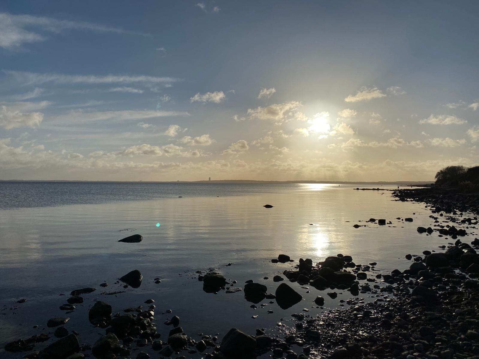 Sonnenlicht reflektiert auf ruhigem Wasser neben einem steinigen Ufer.