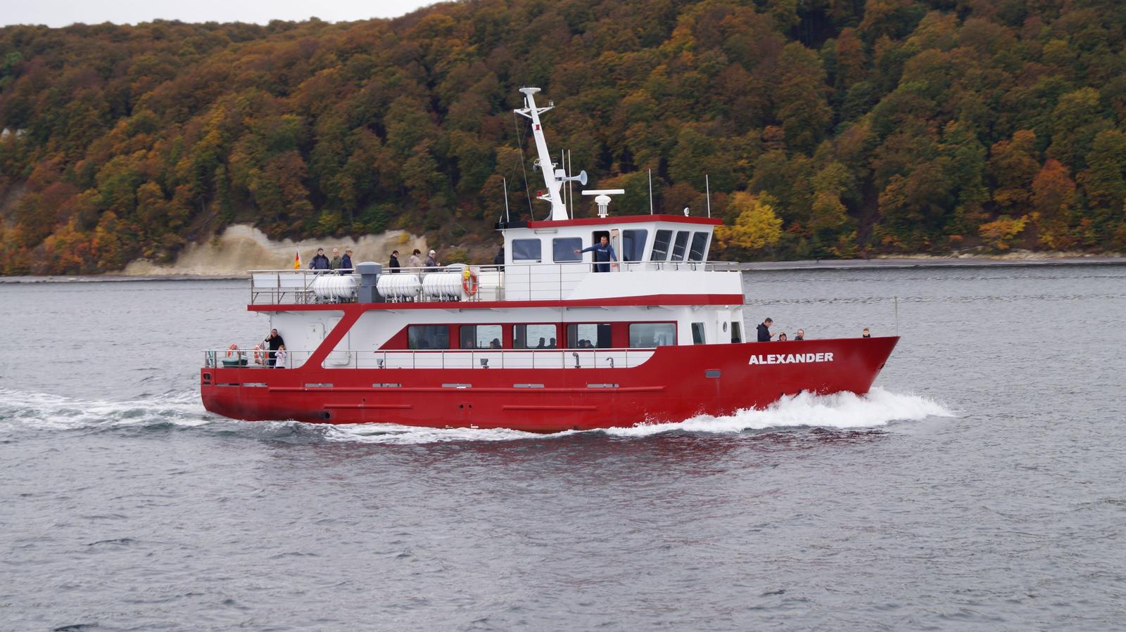 Rotes Schiff mit weißen Aufbauten fährt auf dem Wasser. Menschen stehen auf dem Deck.