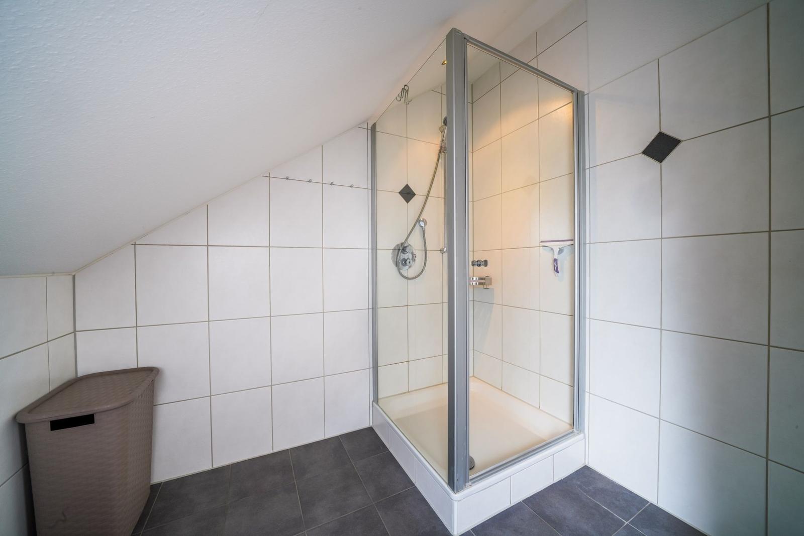 Dusche mit Glaswand, weiße Fliesen, dunkler Boden und Abfalleimer.