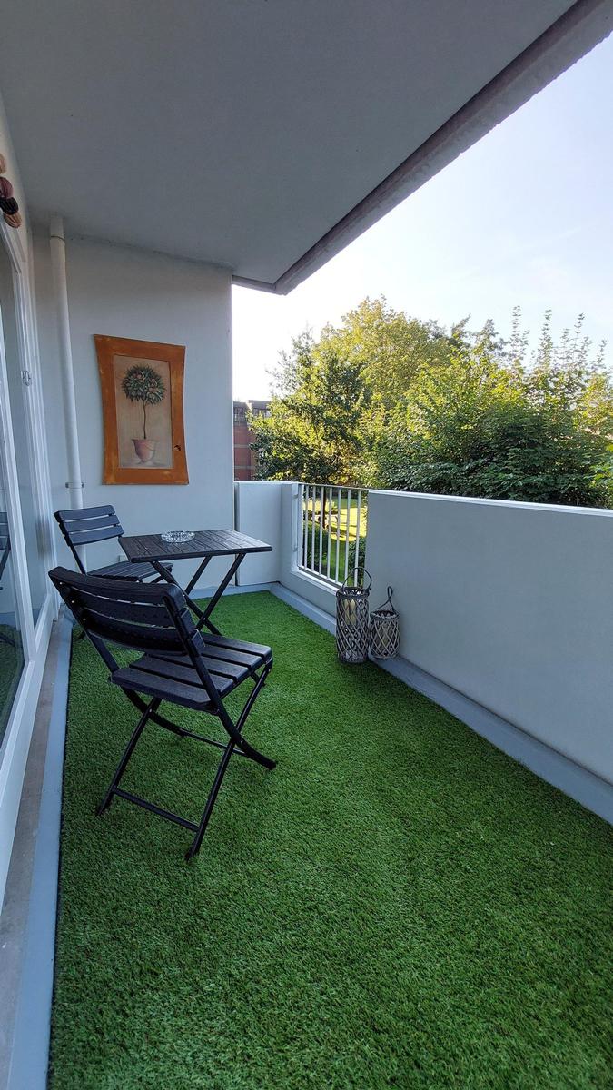 Terrasse mit Tisch, zwei Stühlen und künstlichem Gras. Wandbild und Laternen sind vorhanden.