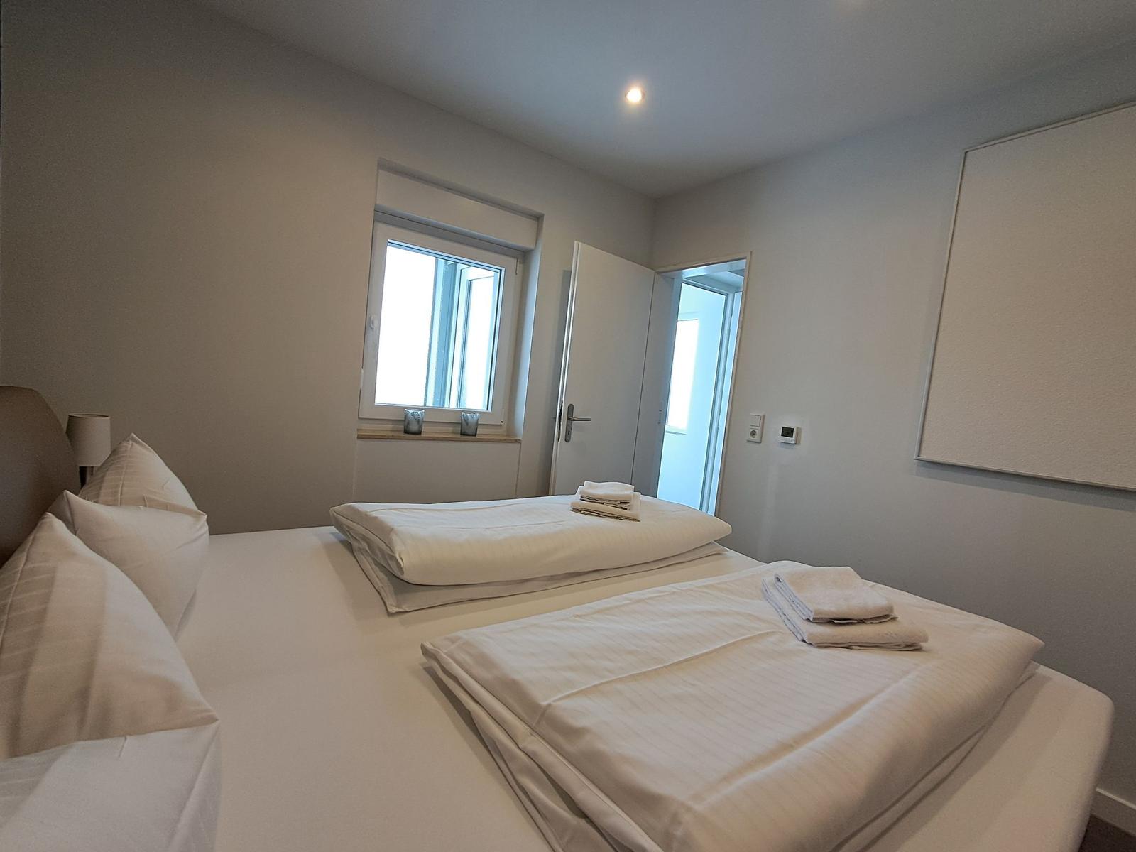 Zimmer mit Bett, Fenster und Tür. Bett mit Bettwäsche und Handtüchern.