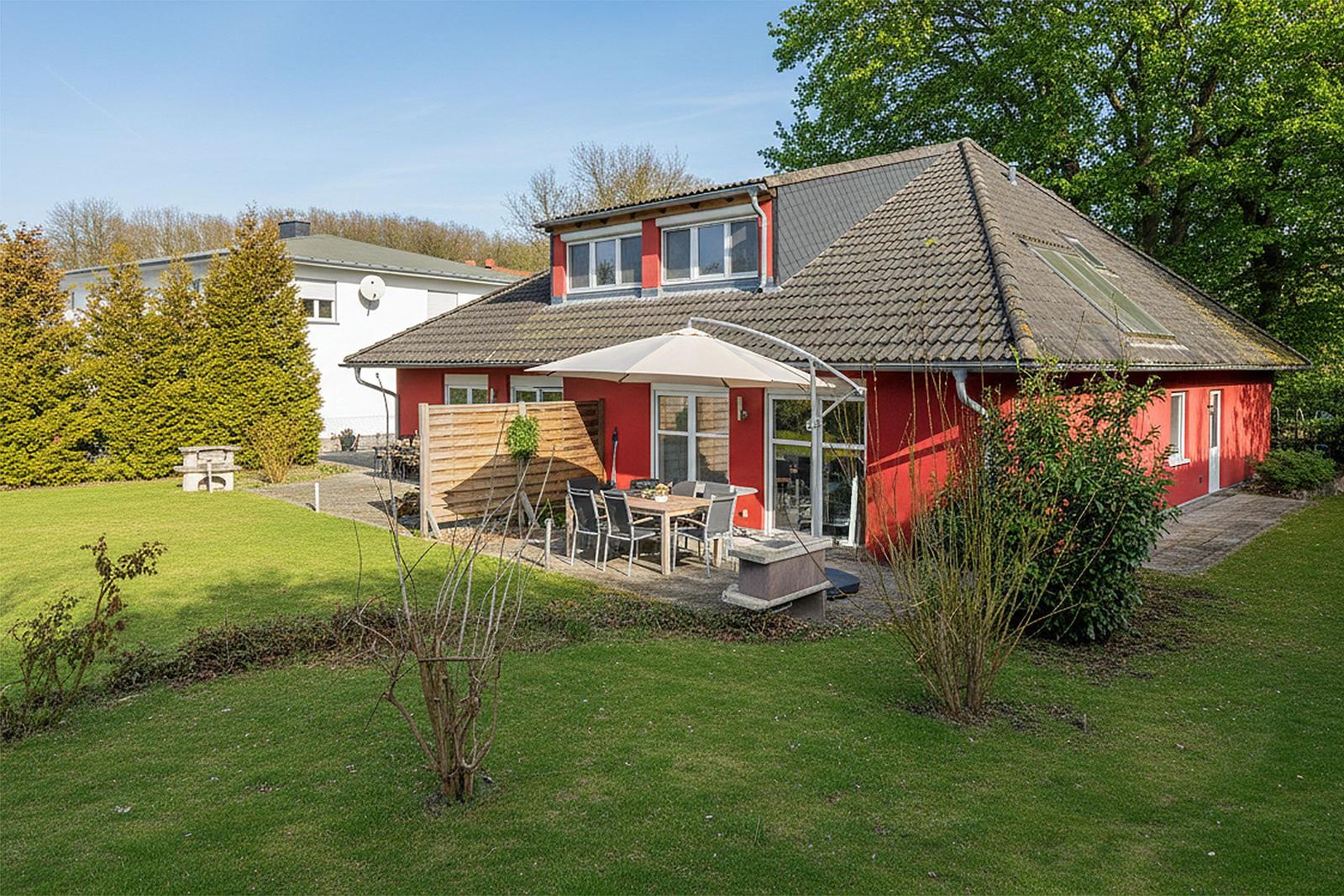 Rotes Haus mit Garten, Terrasse und Sonnenschutz. Grünflächen und Bäume umgeben die Außenanlage.