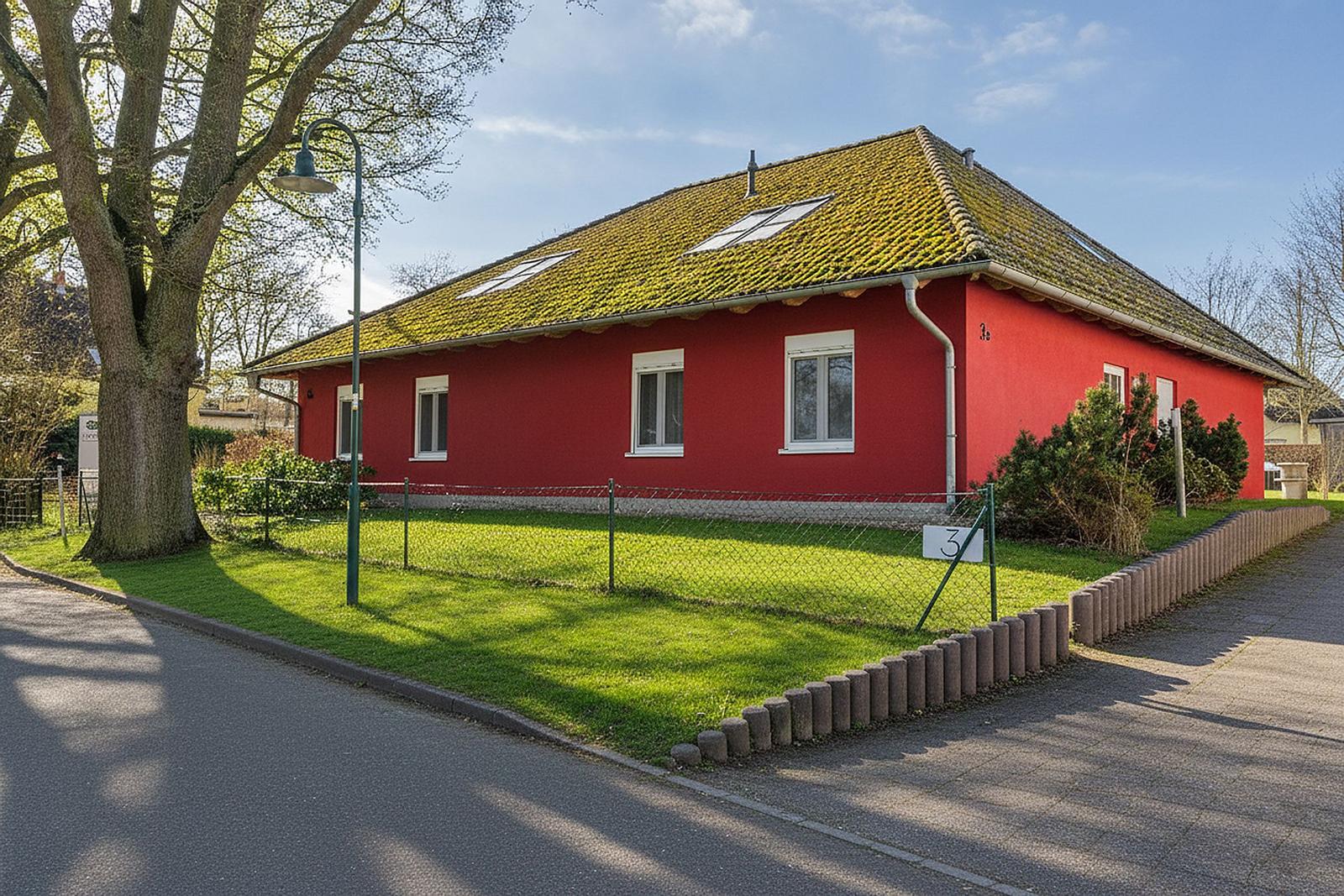 Rotes Haus mit grünem Dach und Vorgarten. Straße und Baum sind sichtbar.