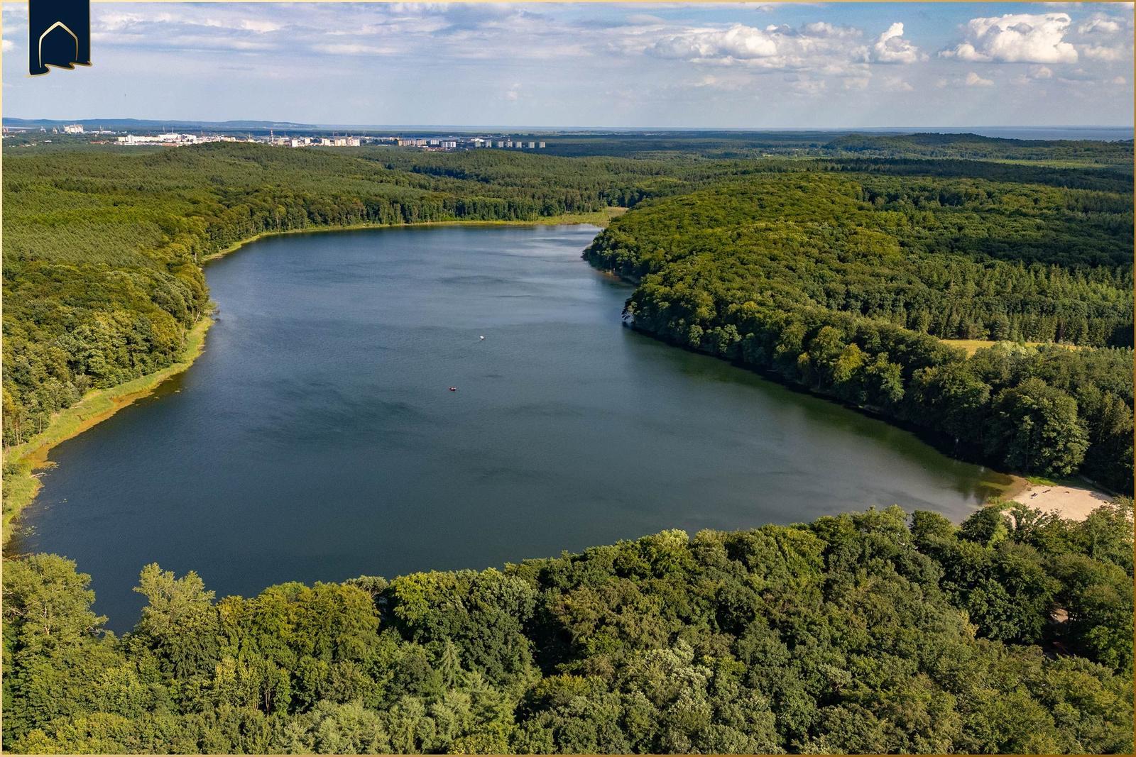 Ein See umgeben von dichtem Wald. Im Hintergrund sind Gebäude sichtbar.