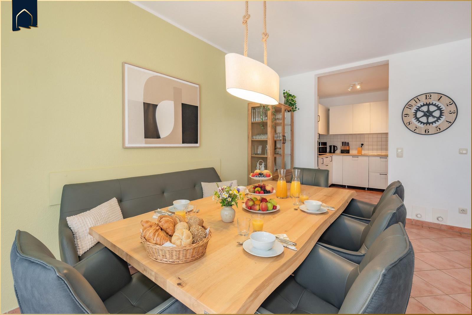 Essbereich mit Holztisch, grauen Stühlen und Frühstücksausstattung. Küche im Hintergrund.