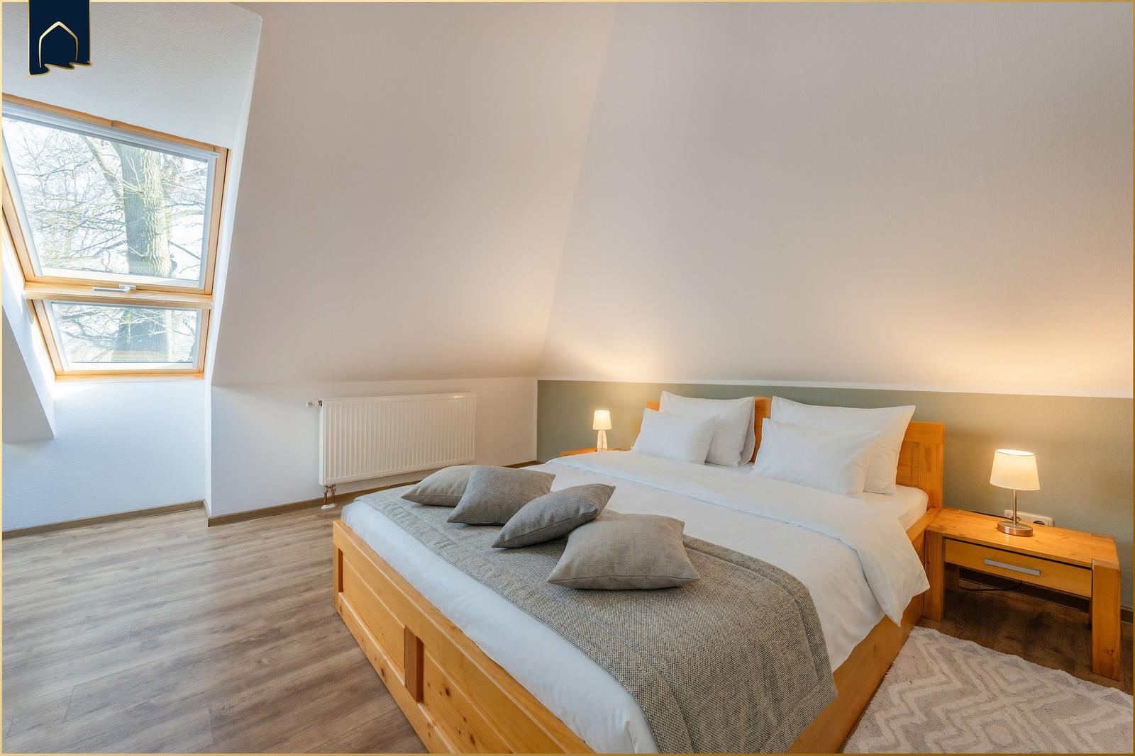 Schlafzimmer mit Dachschrägen, Holzbett, Nachttisch und Fenster mit Blick auf Bäume.